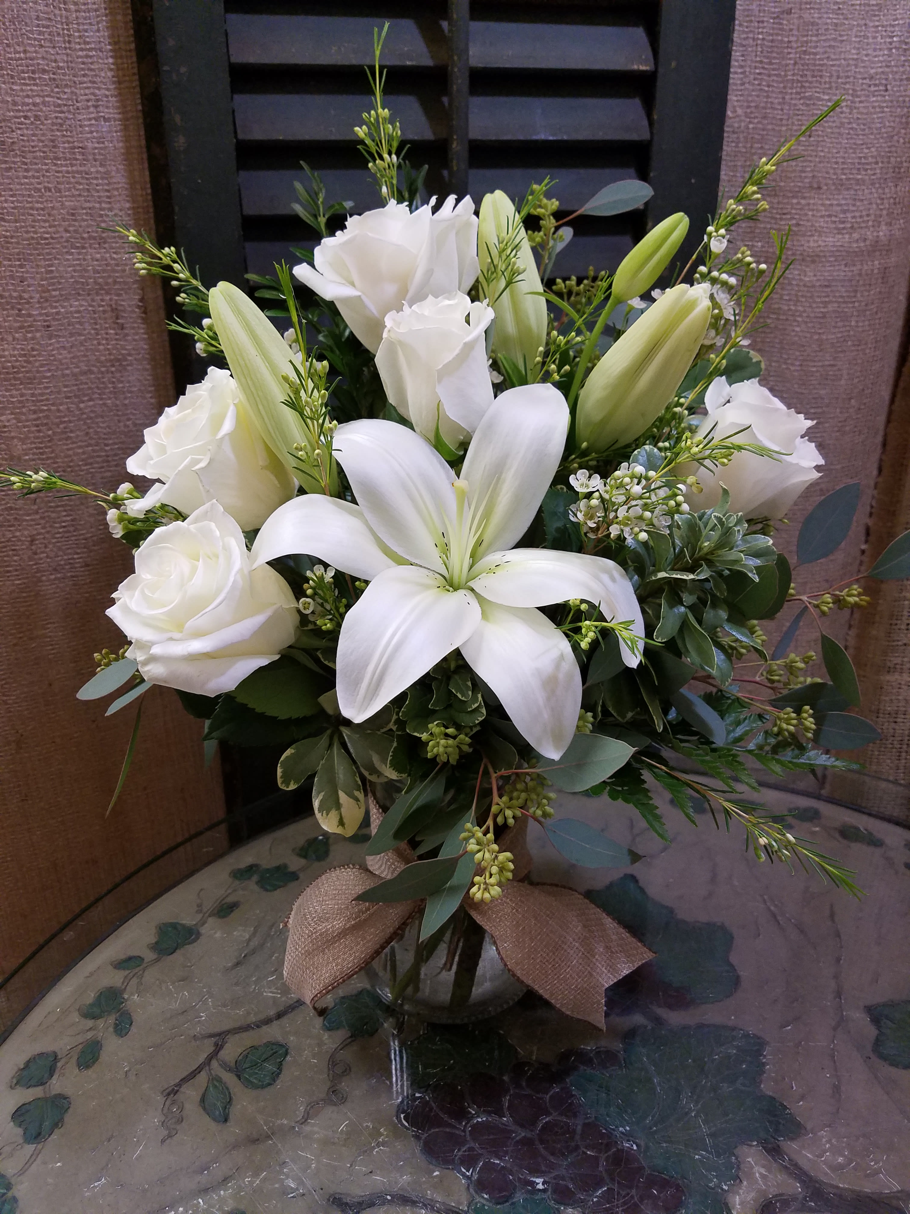 Arrangement of white lilies and roses in a clear glass vase with a burlap bow.