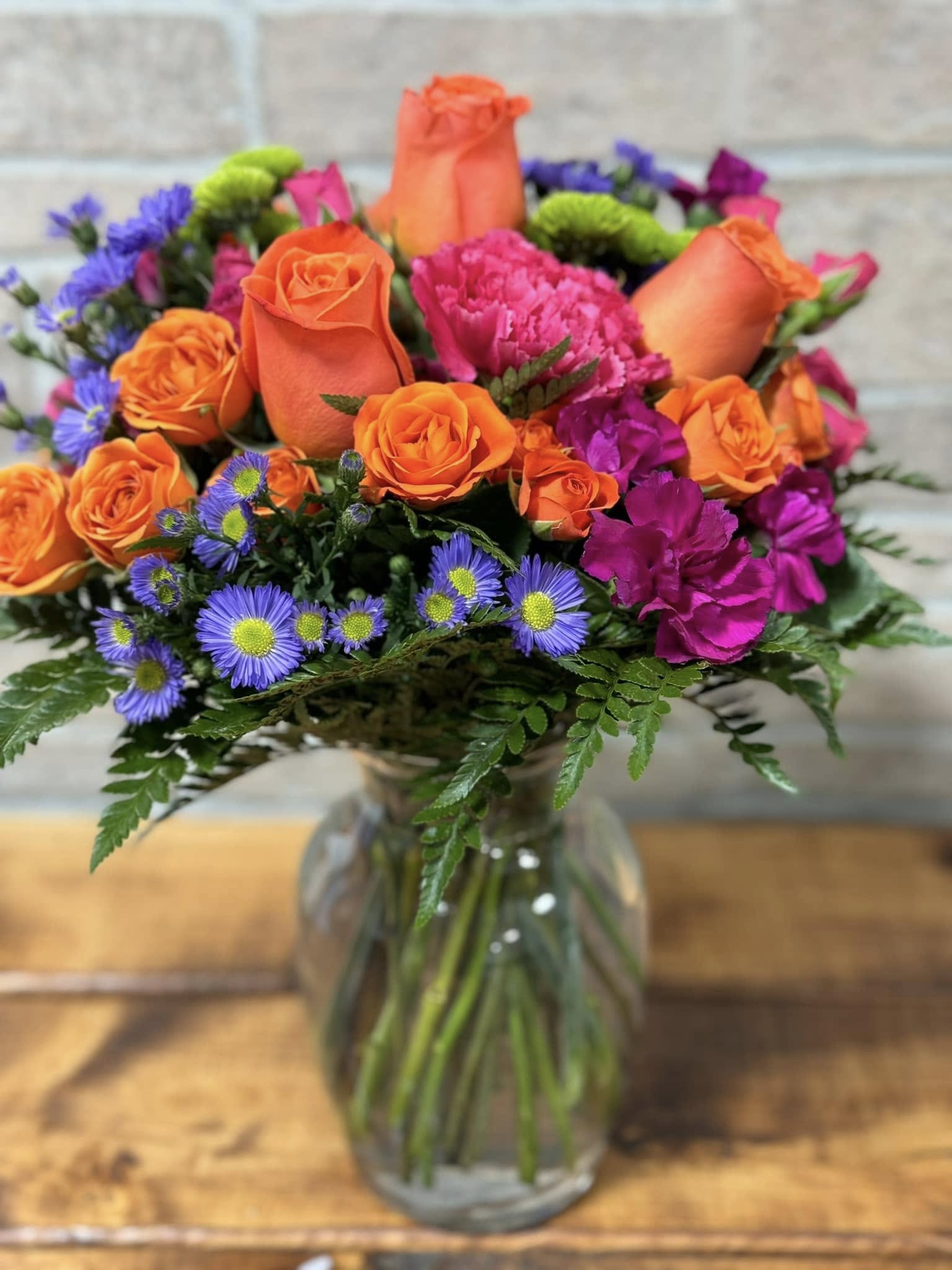 Bright bouquet of orange roses, pink carnations, and purple asters in a clear glass vase