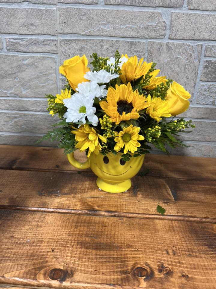Yellow and white flowers arranged in a yellow smiley-face mug on a wooden surface.