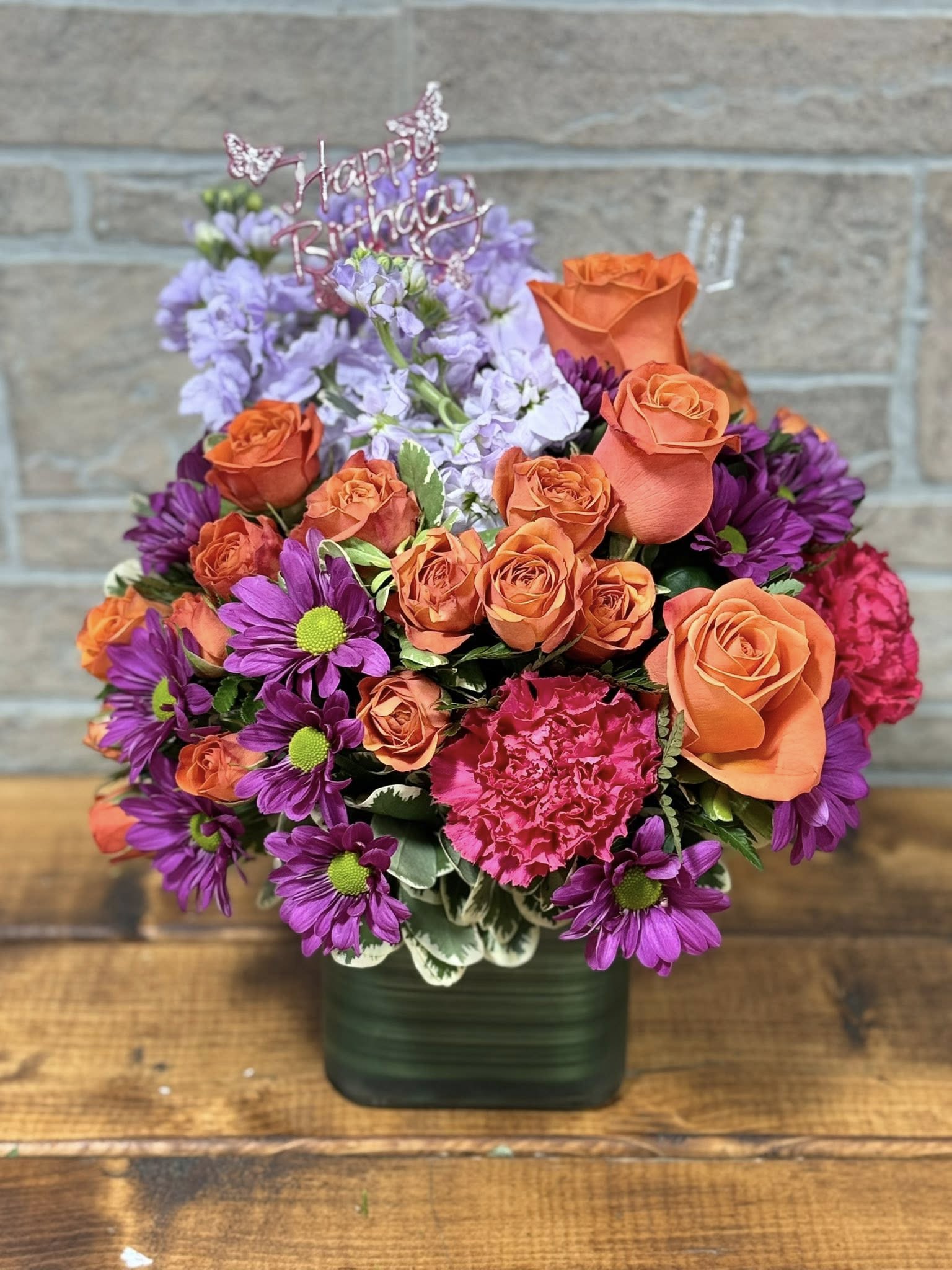 Bright arrangement of orange roses, purple daisies, and pink carnations in a green cube vase with Happy Birthday pick