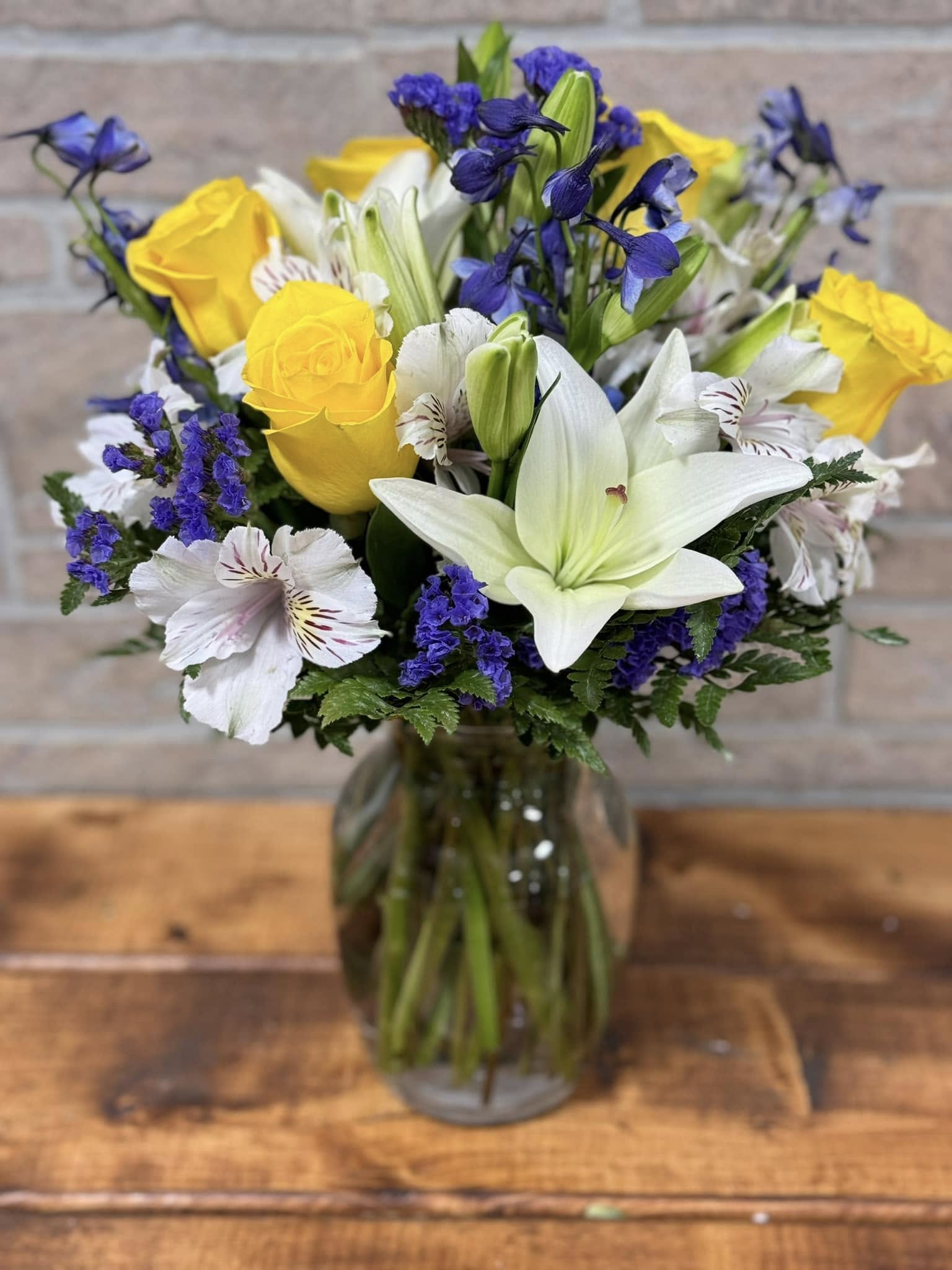 Mixed bouquet of yellow roses, white lilies, and purple flowers in a clear glass vase.