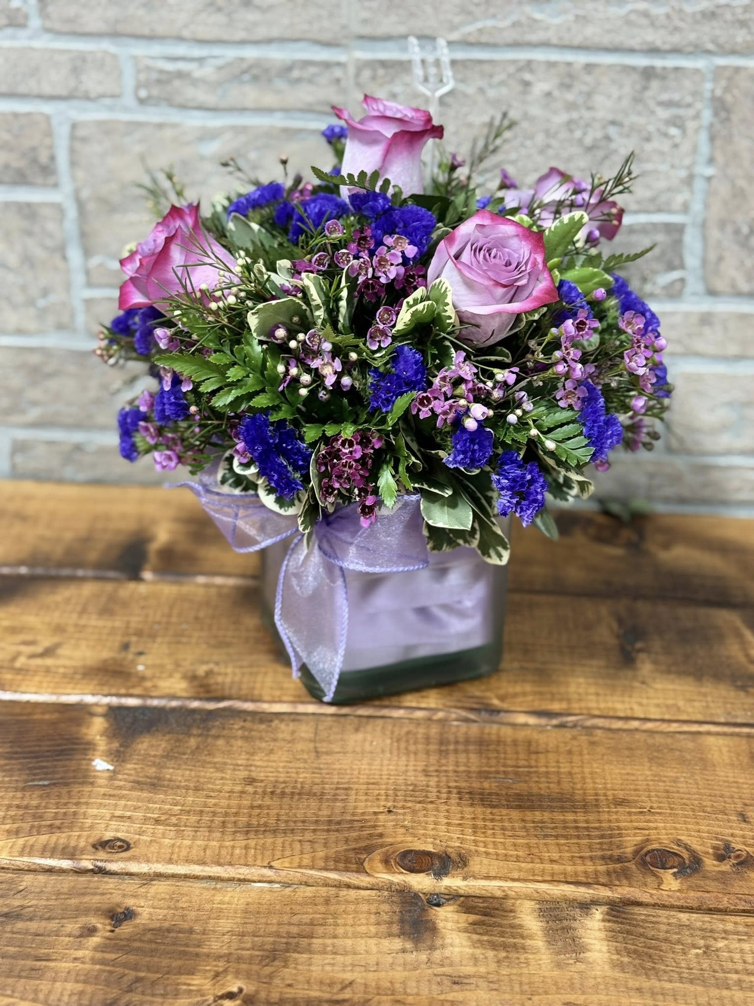 Compact bouquet of pink roses and purple flowers in a square glass vase with a lavender ribbon bow.