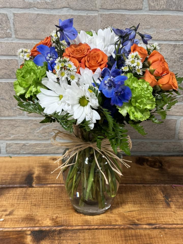 Mixed bouquet of orange roses, blue blooms, white daisies and green carnations in a glass vase with raffia bow