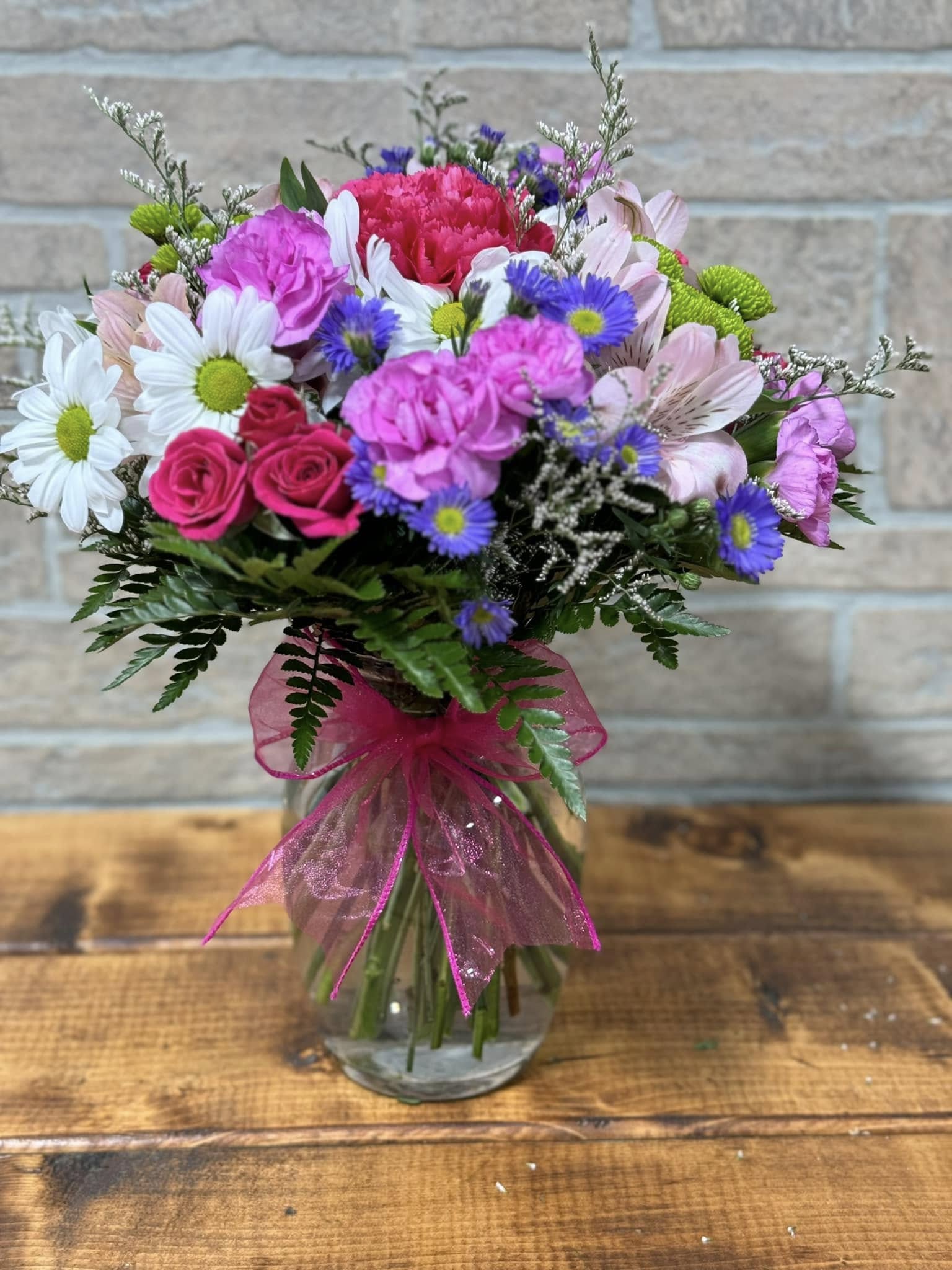 Mixed bouquet of pink, white, and purple flowers in a clear glass vase with a bright pink bow