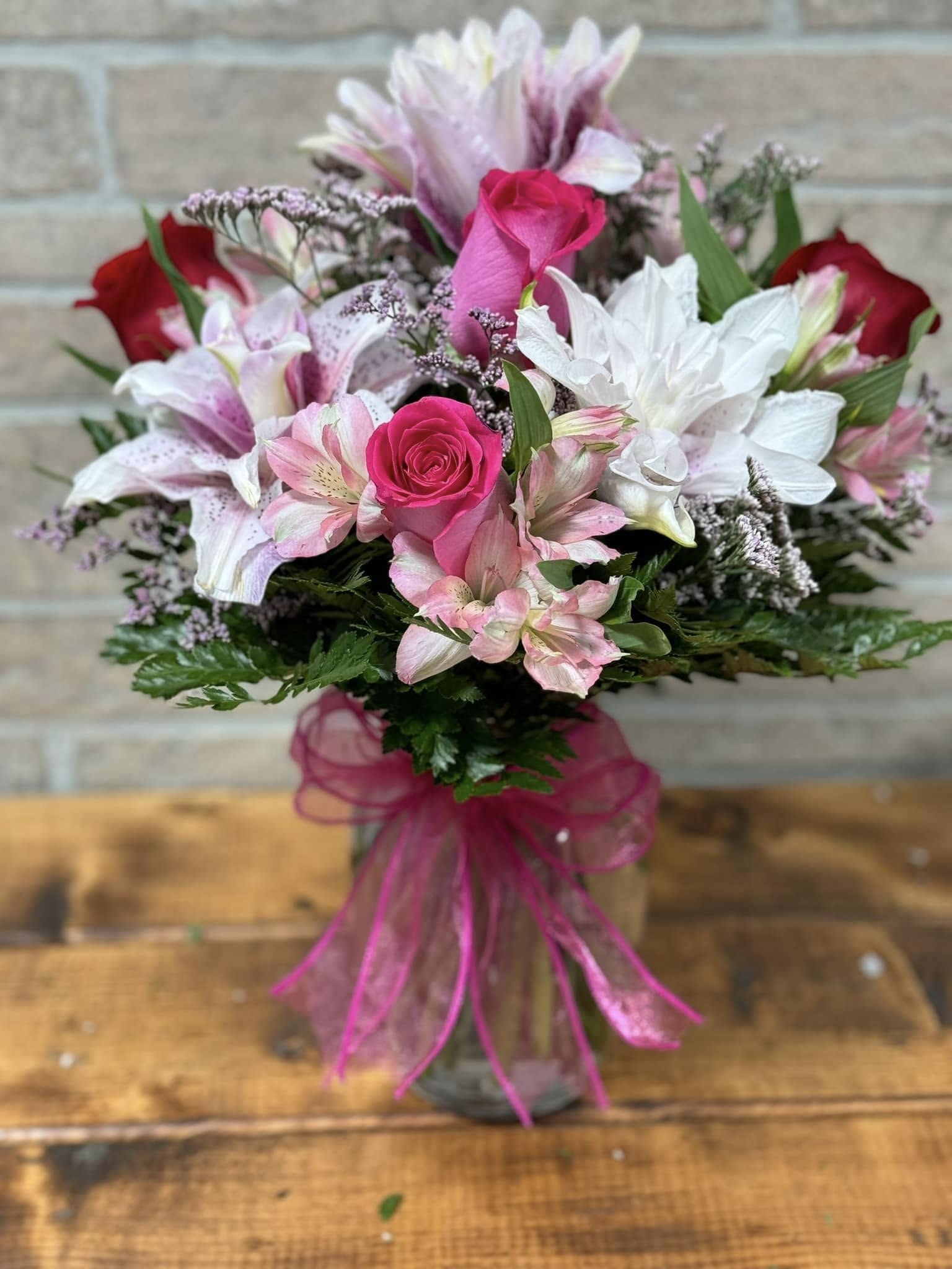 Pink and red roses with white and pink lilies in a clear glass vase with a pink bow.
