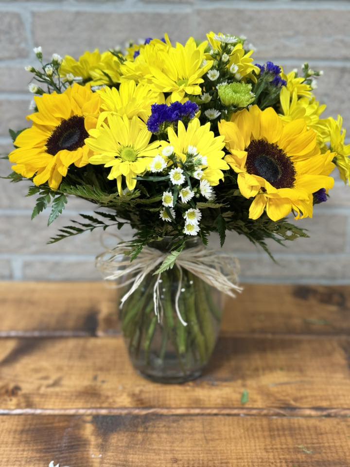 Vase arrangement of bright yellow sunflowers and daisies with white and purple accents.