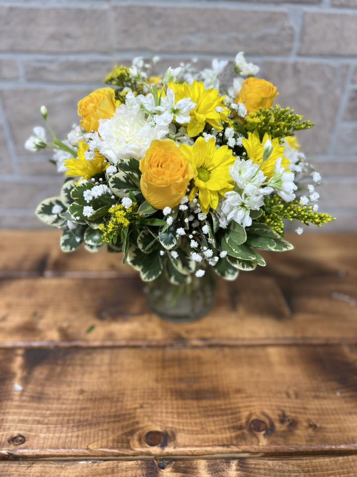 Yellow and white flower arrangement with roses and daisies in a clear glass vase on a wooden table.