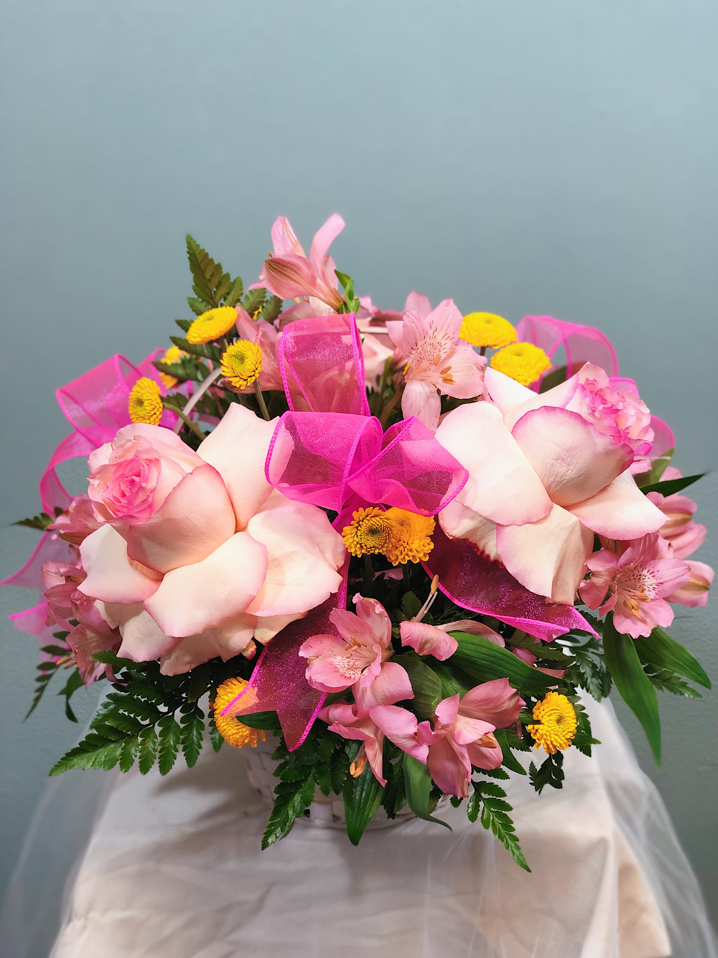 Pink and yellow mixed flower arrangement with roses and alstroemeria in a white basket with bright pink ribbon bows