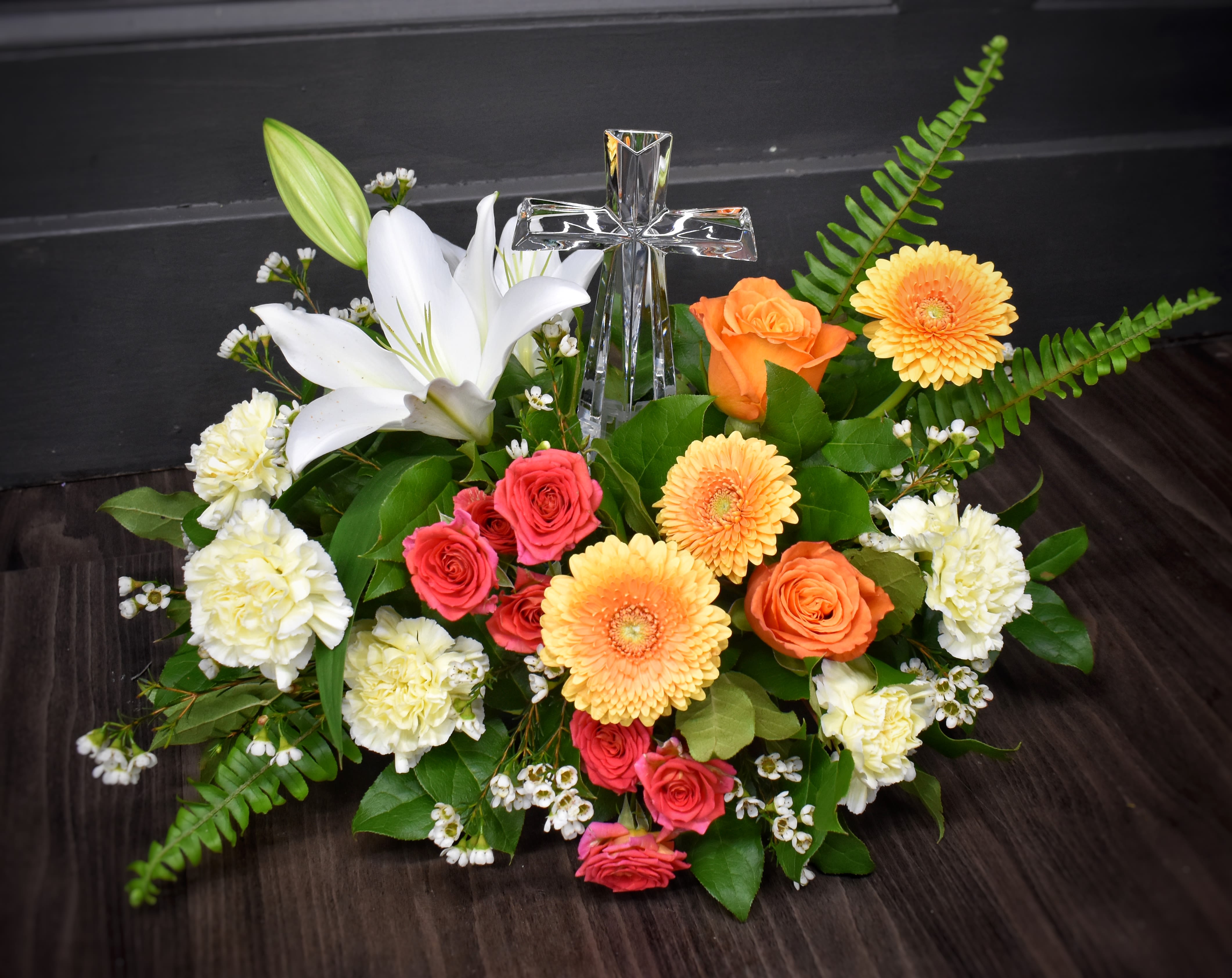 Low arrangement of white lily, peach gerberas, orange and pink roses with a clear glass cross in the center.