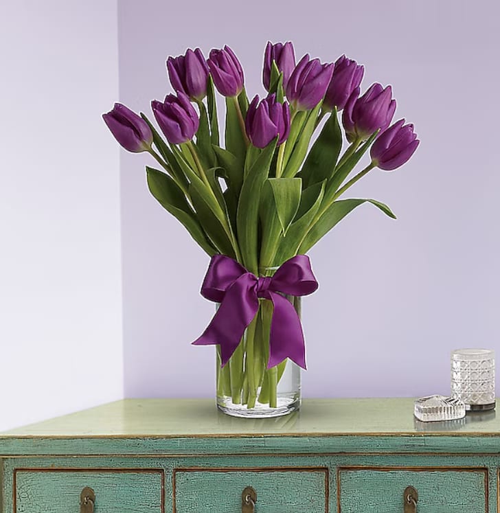 Tall bouquet of purple tulips in a clear glass vase with a matching purple ribbon, set on a green console table.