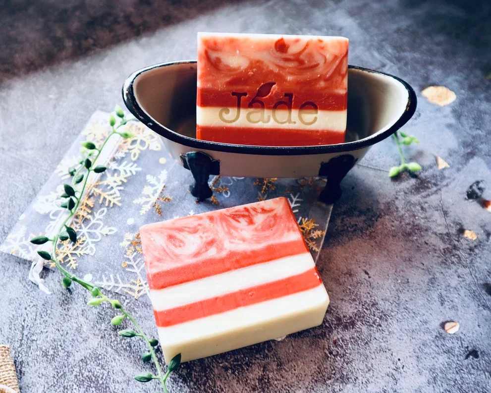 Striped red and white bar soaps, one in a small metal bathtub dish on a textured surface.