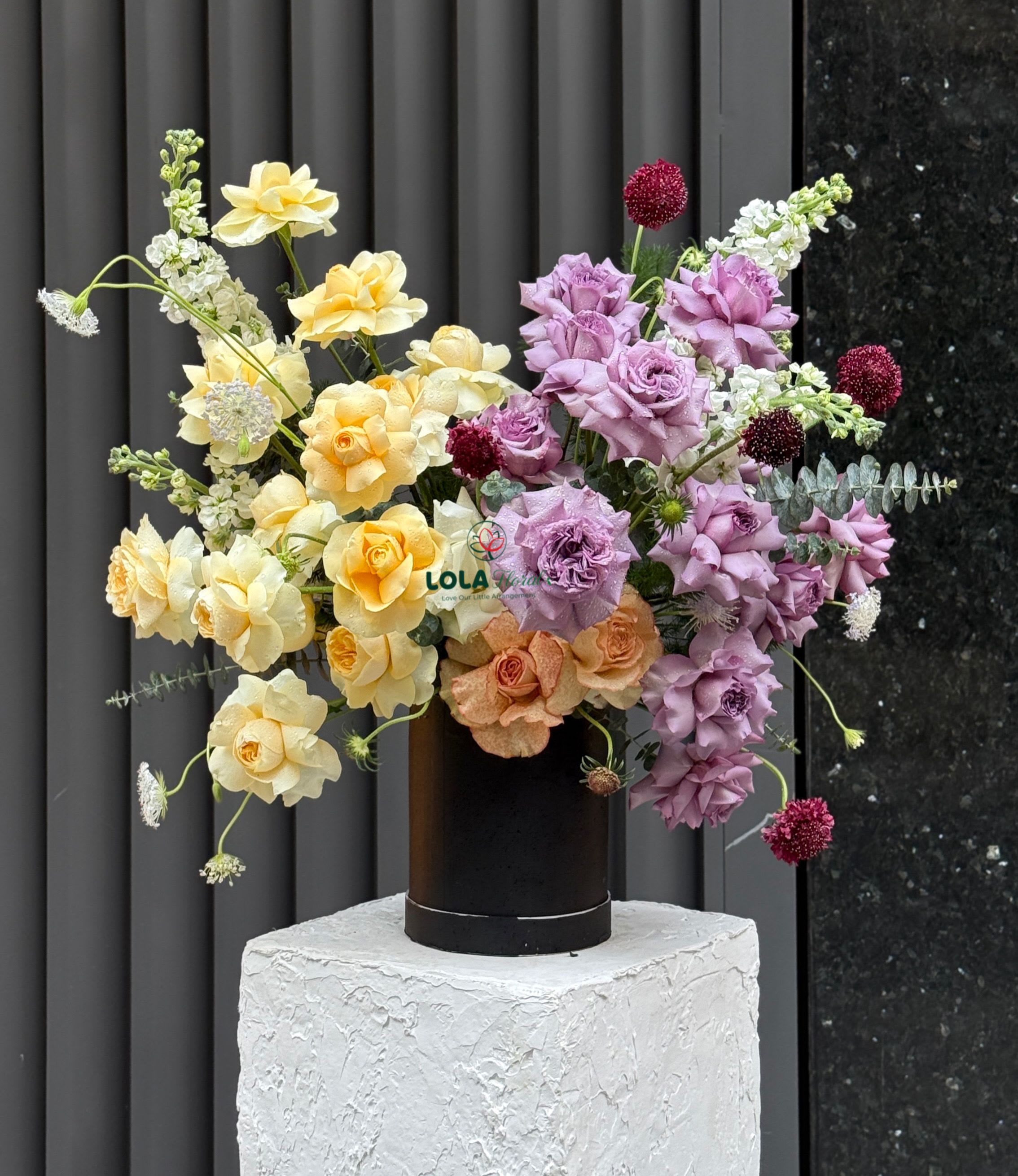 Yellow Purple Vase - Soft yellow and peach roses mingle with lavender roses and burgundy button blooms in this Yellow Purple vase arrangement, a bright, contemporary choice for a birthday bouquet, anniversary flowers, or a thoughtful congratulations gift. Tall white flower spikes and delicate white filler lift the composition into an airy, asymmetrical silhouette that feels both celebratory and refined-perfect for making the person you're thinking of feel genuinely seen and cheered.  The arrangement is gathered in a sleek black cylinder vase and presented atop a textured white pedestal for a gallery-like display that suits modern homes and office settings. Expect a mix of pastel tones-soft yellow, peach, and lavender-accented by deep burgundy spheres and crisp white spikes that add height and contrast. The overall effect is light, textural, and joyful without being fussy.  Ideal for sending to someone marking a milestone or to brighten a table at a small celebration, this arrangement offers visual impact and easy-care enjoyment; place it in a cool spot away from direct sunlight and refresh the water every 2-3 days to keep roses looking their best. If you need specific sizing or delivery timing, select your preferred options at checkout to ensure it arrives when the moment matters.
