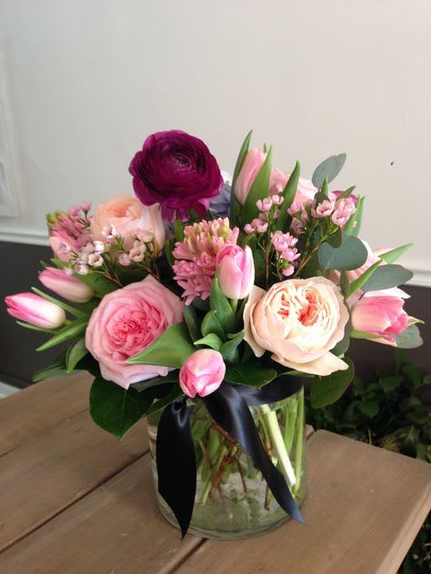 Pink roses, tulips, and mixed blooms in a clear glass vase tied with a black ribbon