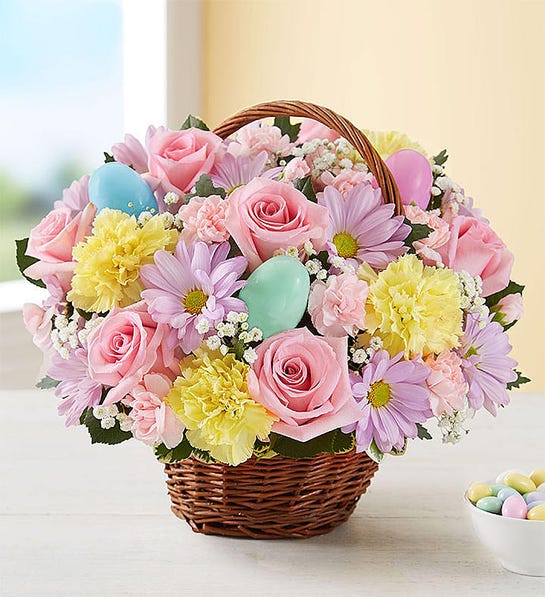 Pastel basket arrangement with pink roses, yellow carnations, lavender daisies, and decorative Easter eggs.