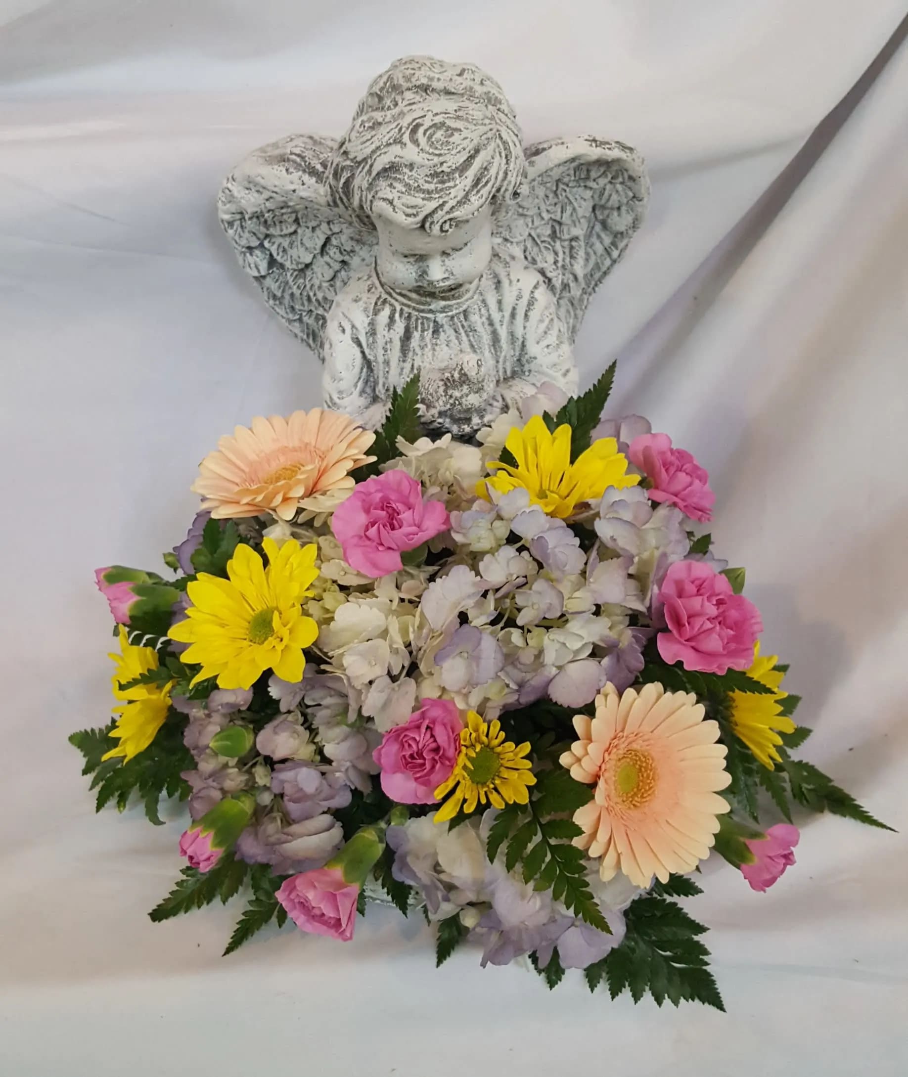 Angel figurine with low pastel flower arrangement of yellow daisies, pink blooms, and hydrangeas at the base.