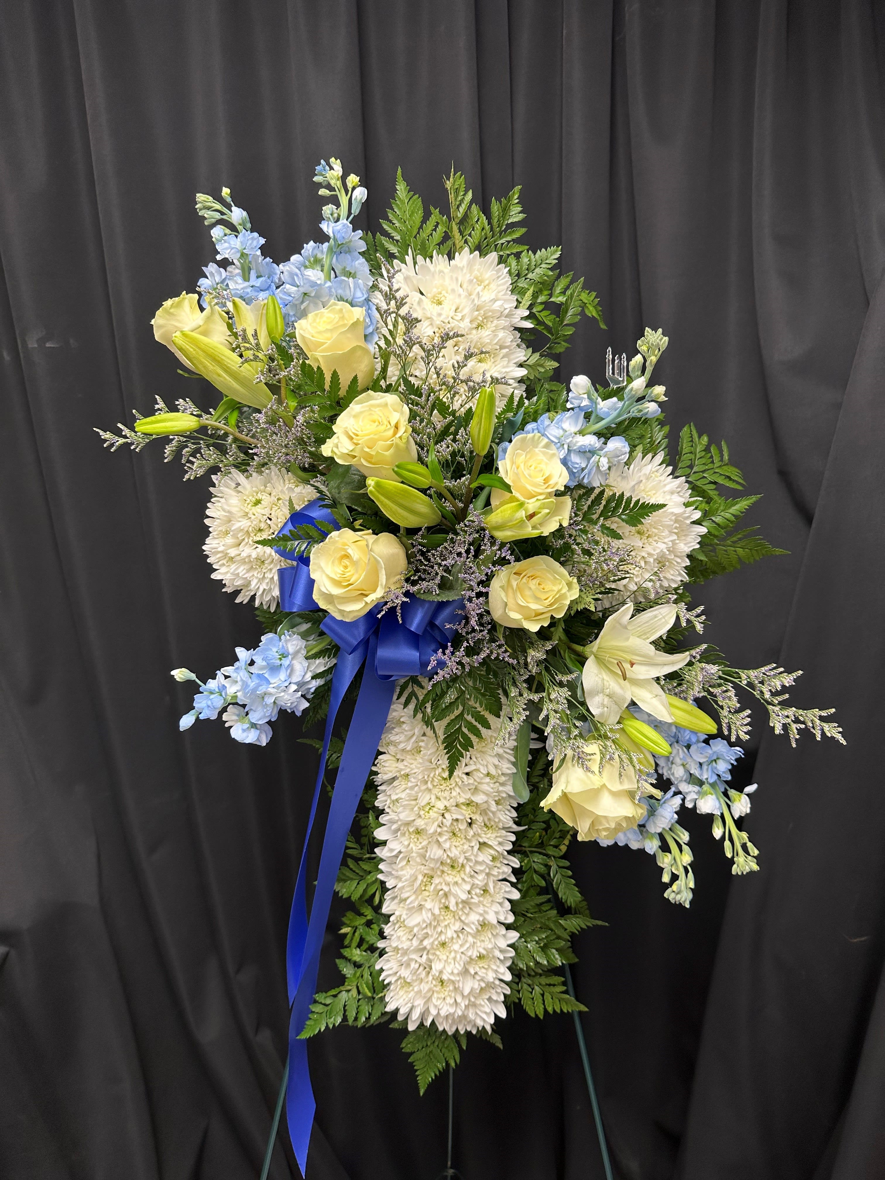Standing spray of white mums, yellow roses, lilies, and blue flowers with blue ribbon on an easel.