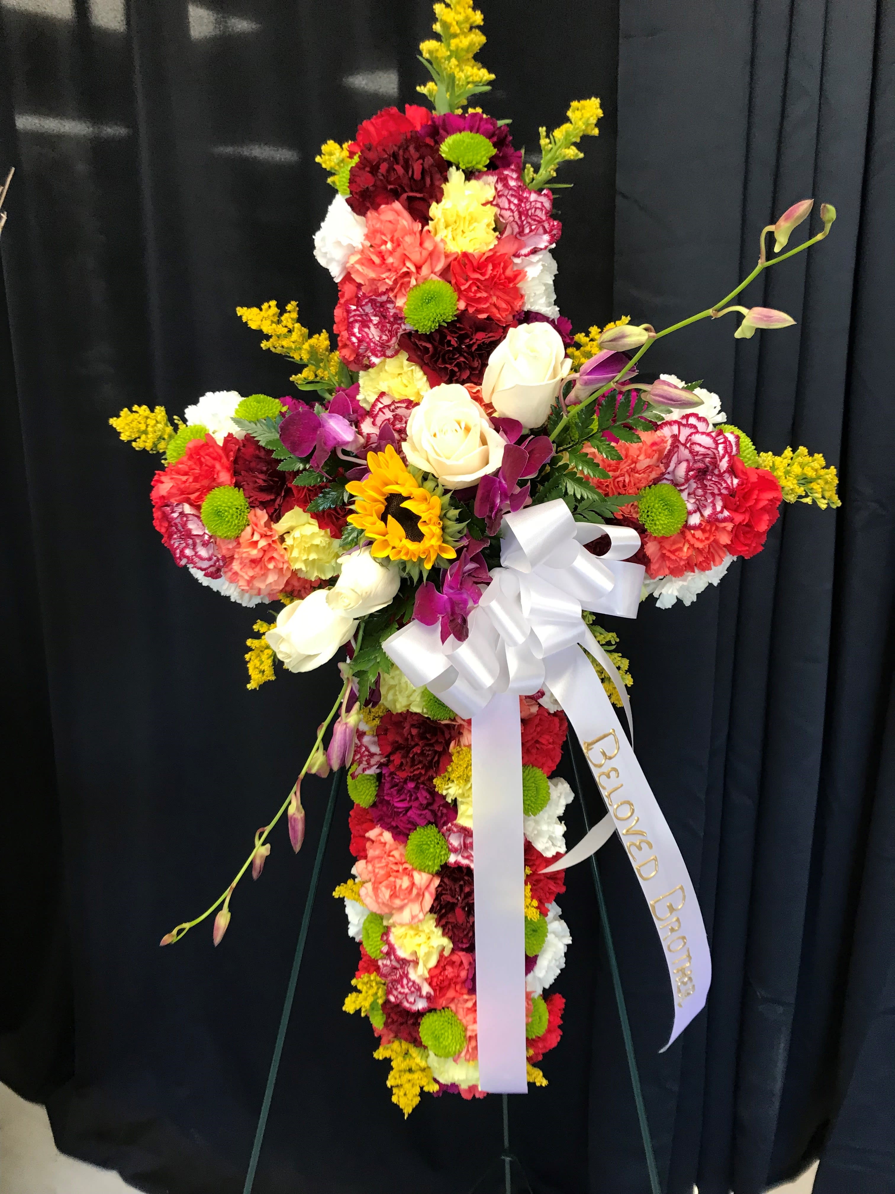 Standing cross of mixed bright flowers with roses and sunflower, white bow, and 'Beloved Brother' ribbon.