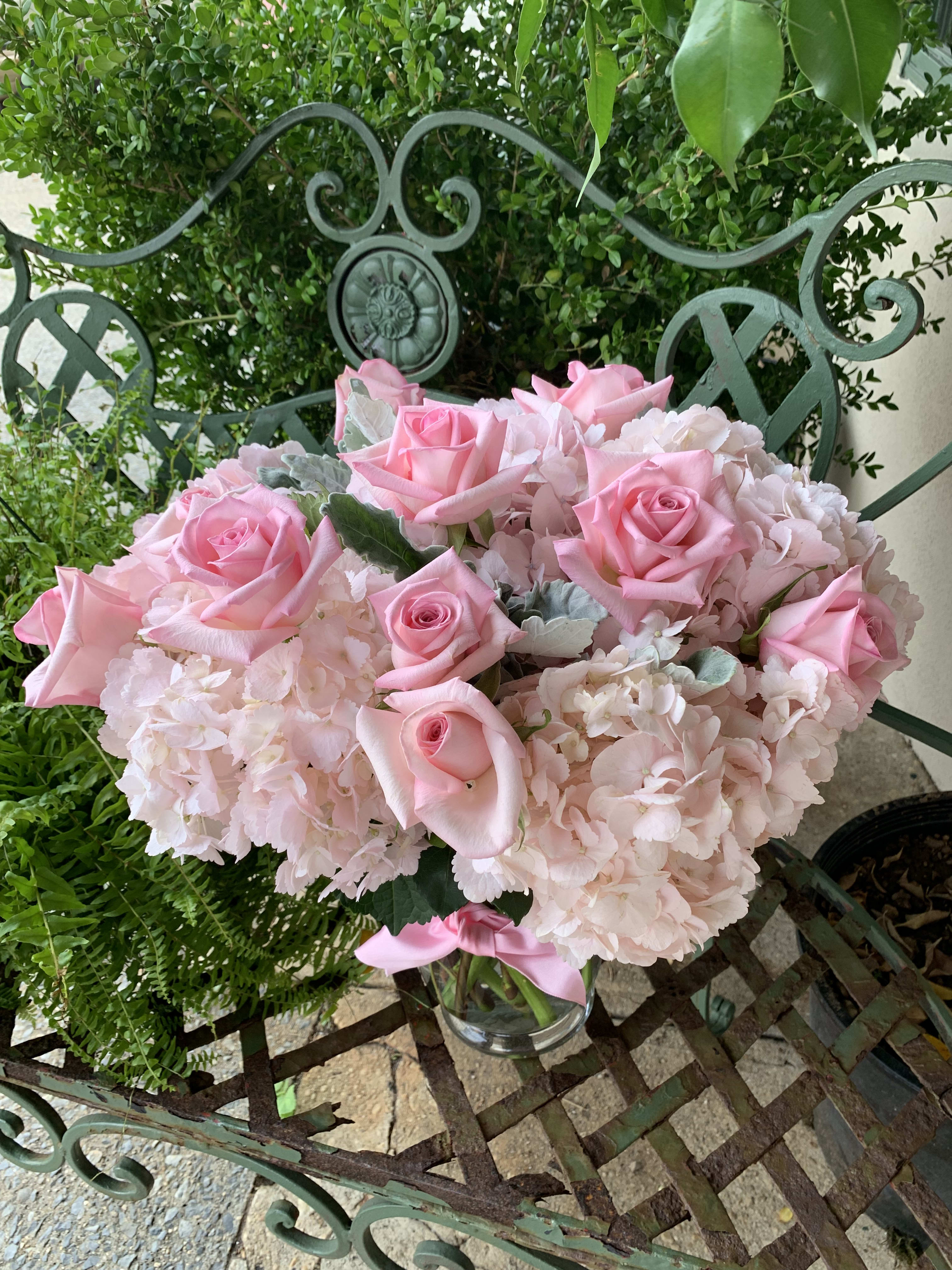 Low glass vase of light pink roses and hydrangeas arranged in a compact mound on a metal bench