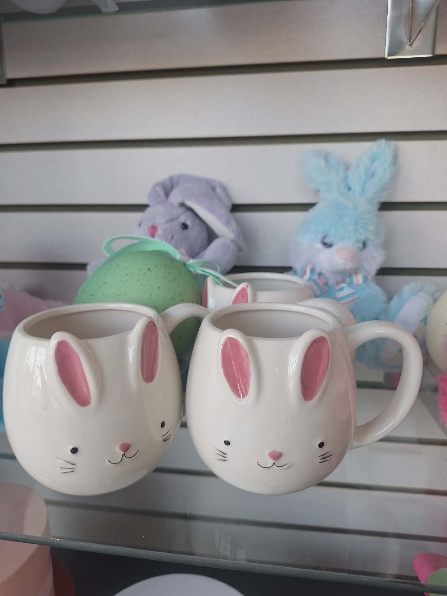 Two white bunny-face mugs with pink ears on a glass shelf with pastel bunny toys behind.
