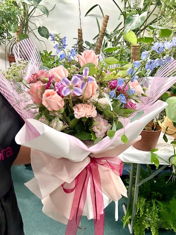 Hand-tied bouquet of peach roses and pastel mixed flowers wrapped in white and pink paper with a magenta ribbon
