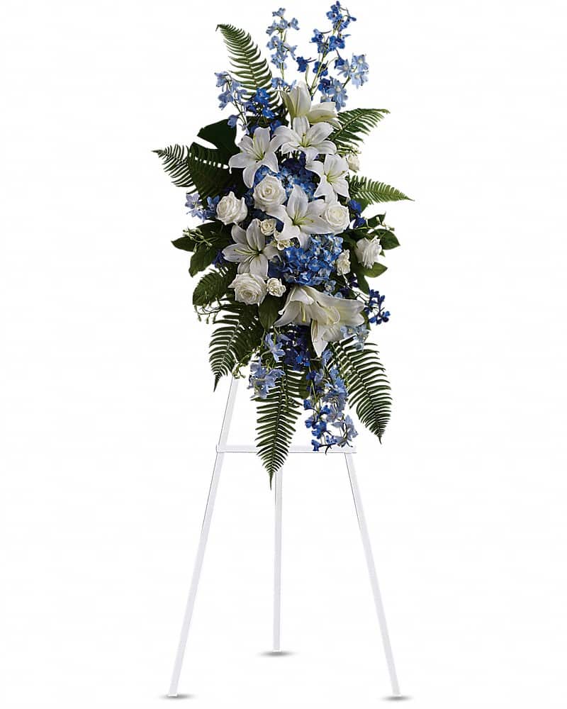 Tall standing spray of white lilies and roses with blue hydrangea and blue delphinium on a white easel
