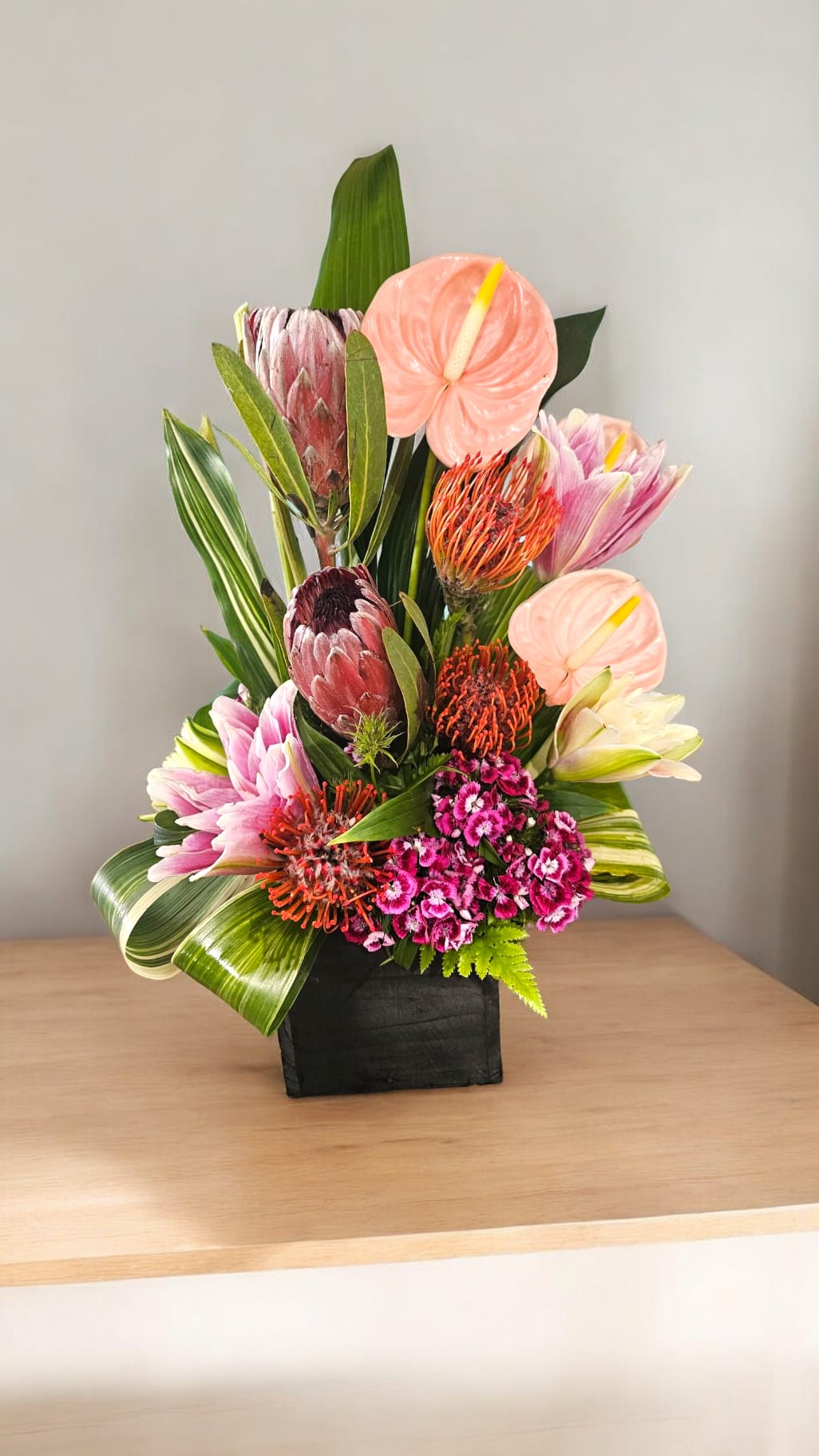 Tropical Radiance - Create a stunning tropical atmosphere with our Tropical Radiance arrangement. Featuring vibrant orange and pink protea, delicate oriental lilies, and hot pink dianthus, all nestled in lush tropical greenery on a stylish wooden cube. Guaranteed to add a touch of exotic elegance to any space.
