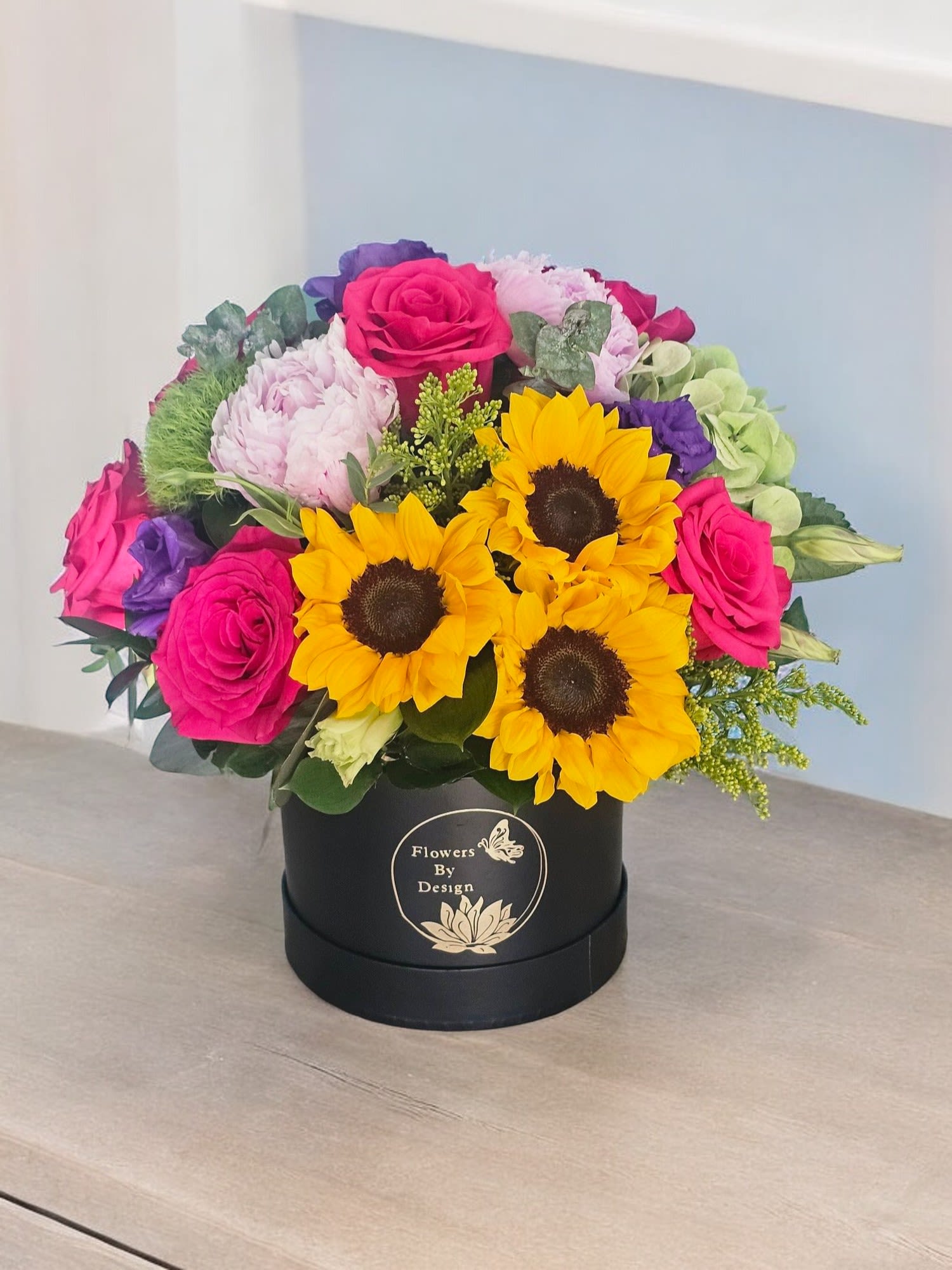 Fuchsia Garden Elegance - For this elegant and vibrant arrangement is made It highlights the bold contrast of hot pink roses against the deep hues of sunflowers and purple lisianthus, complemented by the fresh greens of hydrangeas and trick dianthus, all accented with lush greenery and delicate filler.