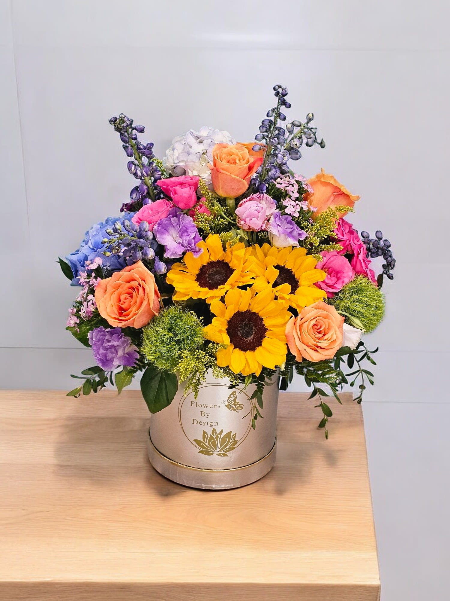 Sunlit Garden Symphony - The essence of the arrangement is made with Premiun Sunflower, Orange Roses, Blue Delphium, Purple Lisianthus, Green Tick, Pink dianthus, Blue Hydreangas, some greenery and fine Filler.  This Arrangement is subject at subtitution on Flowers depending on Availability.
