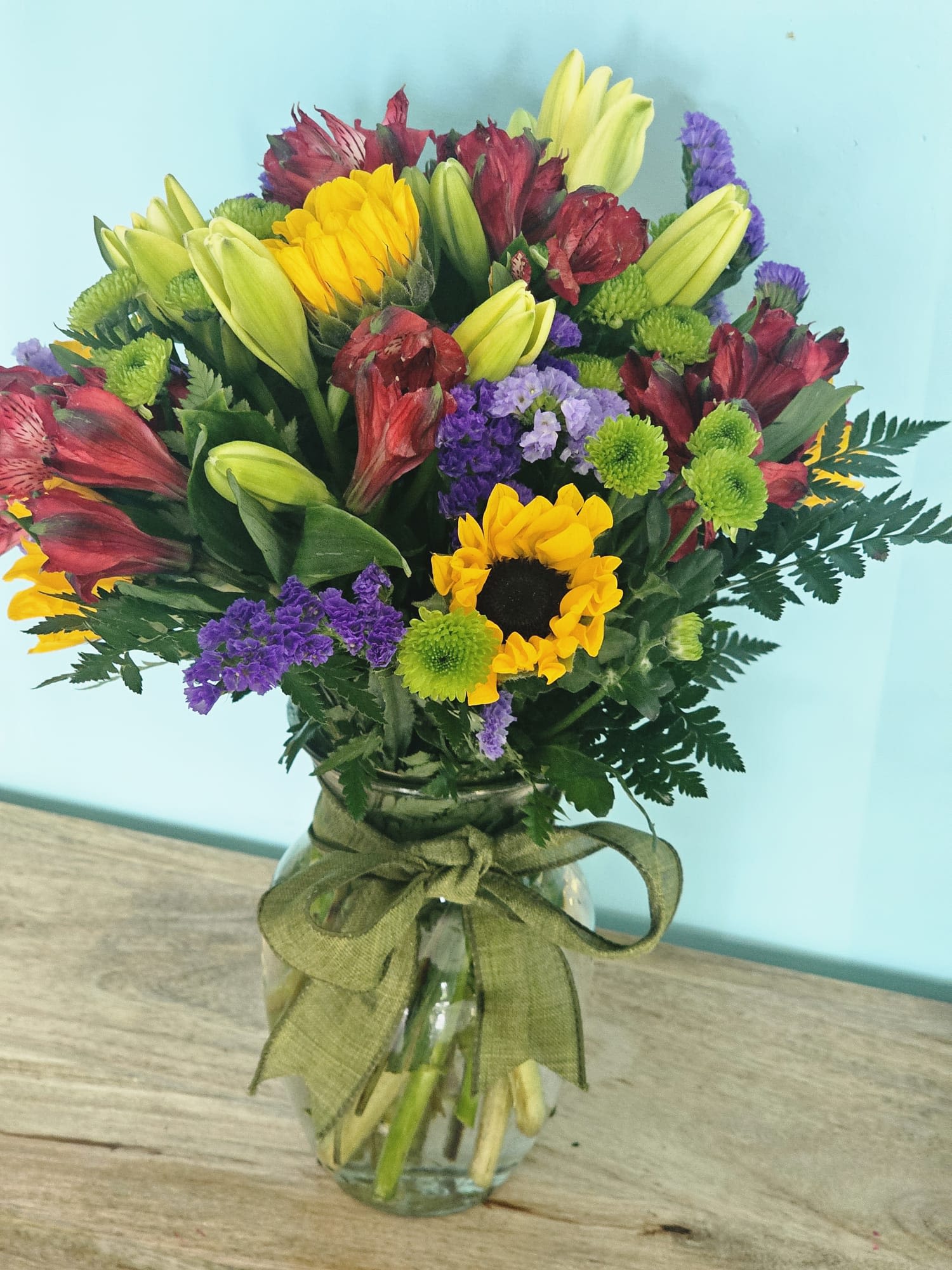 SUMMER TIME - Expertly crafted to evoke the essence of summer, this vibrant arrangement showcases stunning sunflowers, yellow lilies, and red alstroemeria. The composition is enhanced with purple static flowers and lush greenery, all beautifully presented in a clear vase. The contrasting colors create a sophisticated seasonal statement that brings warmth and joy to any space.