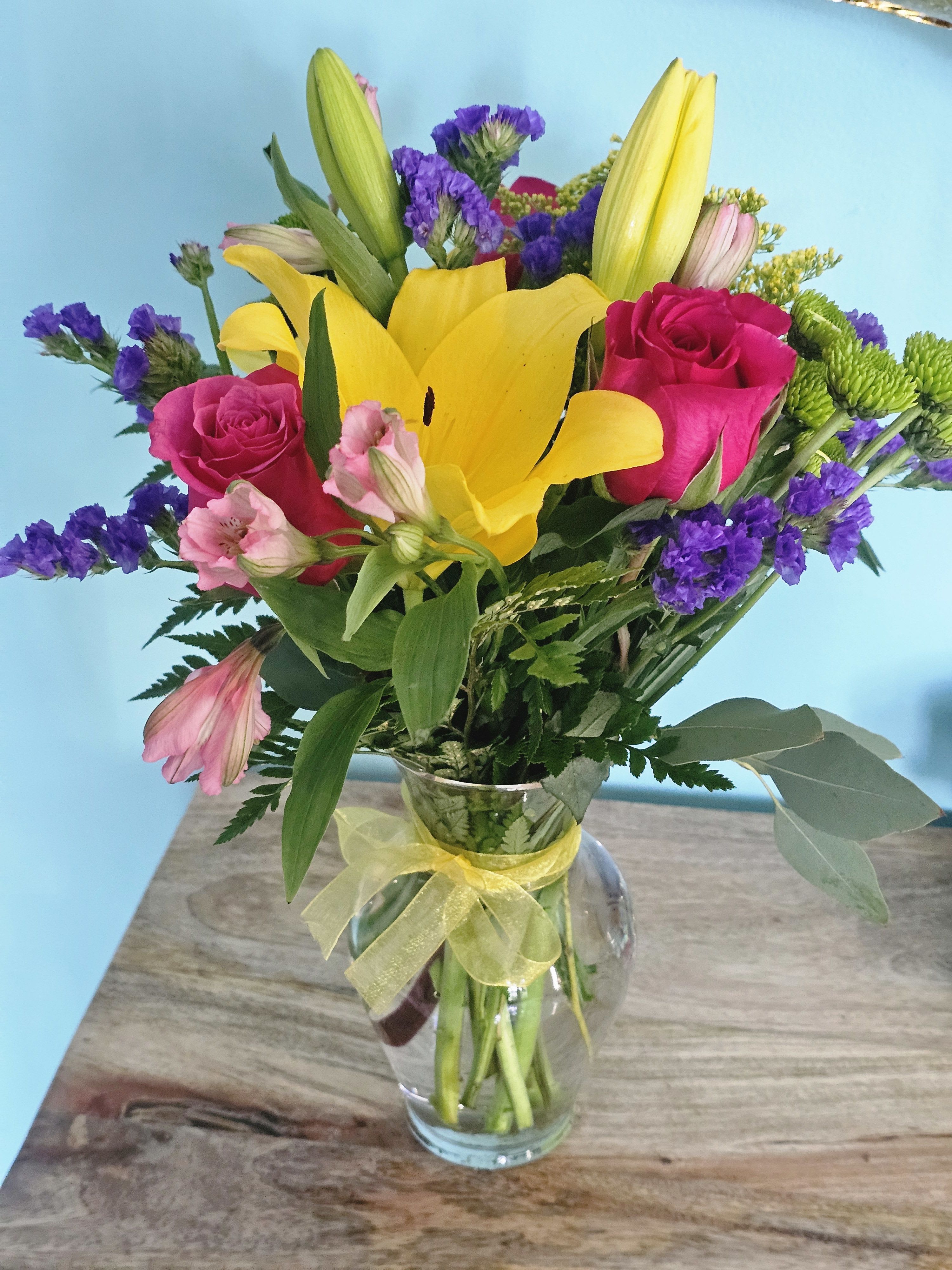 Summer Light - Experience the vibrant beauty of Summer Light with our hot pink roses, yellow lilies, green athos, pink alstromeria, purple static, and solidago fine greenery. Arranged in a clear vase, this bouquet will bring joy and warmth to any space. Perfect for any summer occasion.