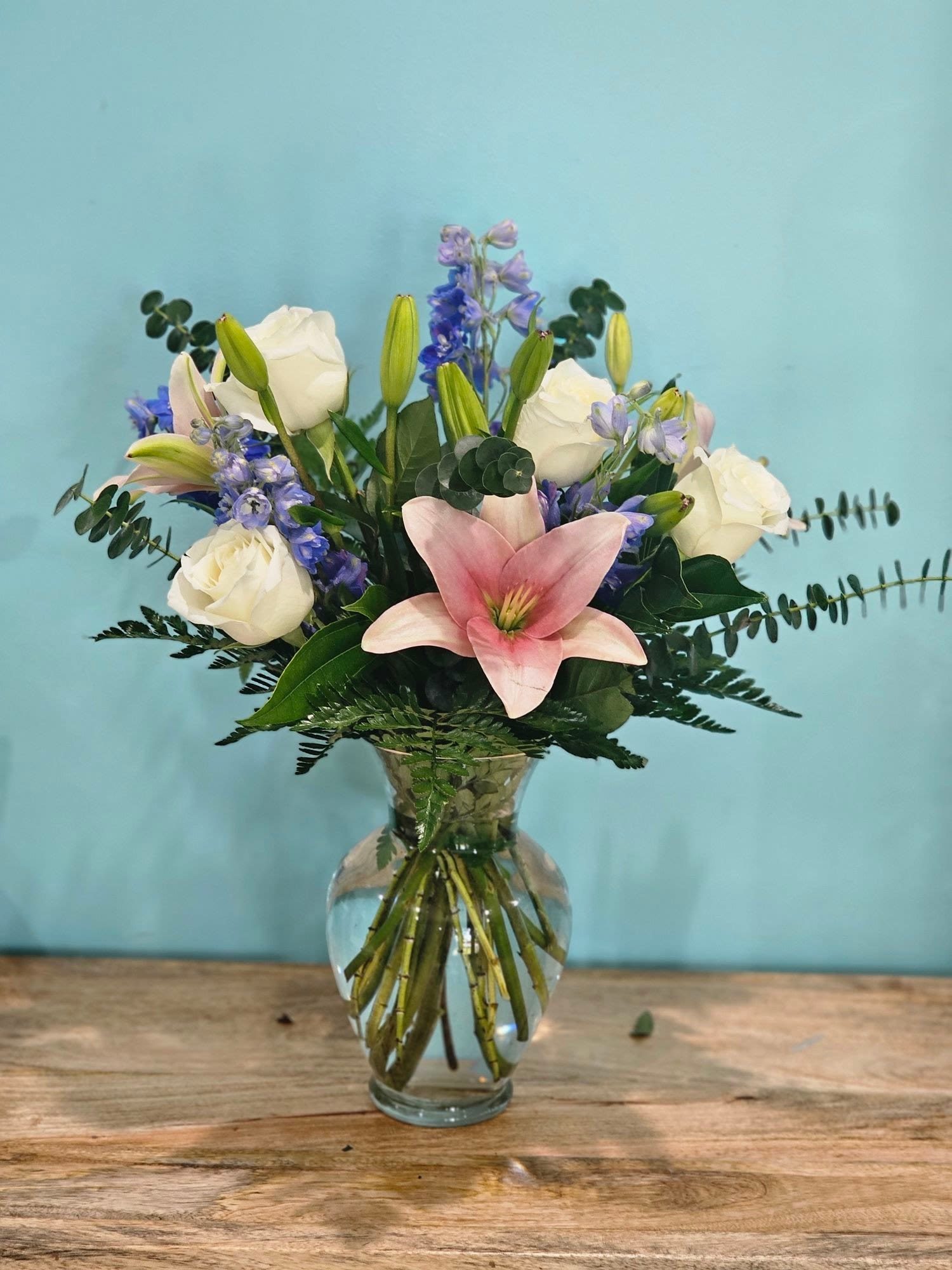 Blissful Blossom Medley - A vase arrangement with white roses, pink lilies, blue delphiniums, and fine greenery sounds absolutely lovely! White roses often symbolize purity and innocence, pink lilies represent prosperity and admiration, and blue delphiniums add a touch of elegance and grace to the mix. The fine greenery will complement these flowers beautifully, providing a lush backdrop and enhancing the overall arrangement. It would make a perfect centerpiece for a variety of occasions, from weddings to special dinners or even as a thoughtful gift