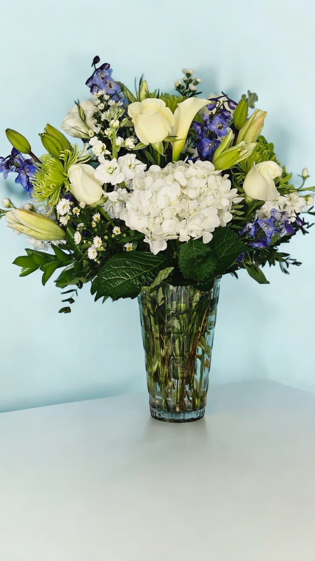 Tranquil Harmony - This Arrangement reflect the soothing and sophisticated combination of colors and flowers, emphasizing the harmonious and elegant nature of the arrangement. An arrangement featuring white callas, blue delphinium, green hydrangeas, white roses, green mums, white stock, white hydrangeas, filler, and greenery