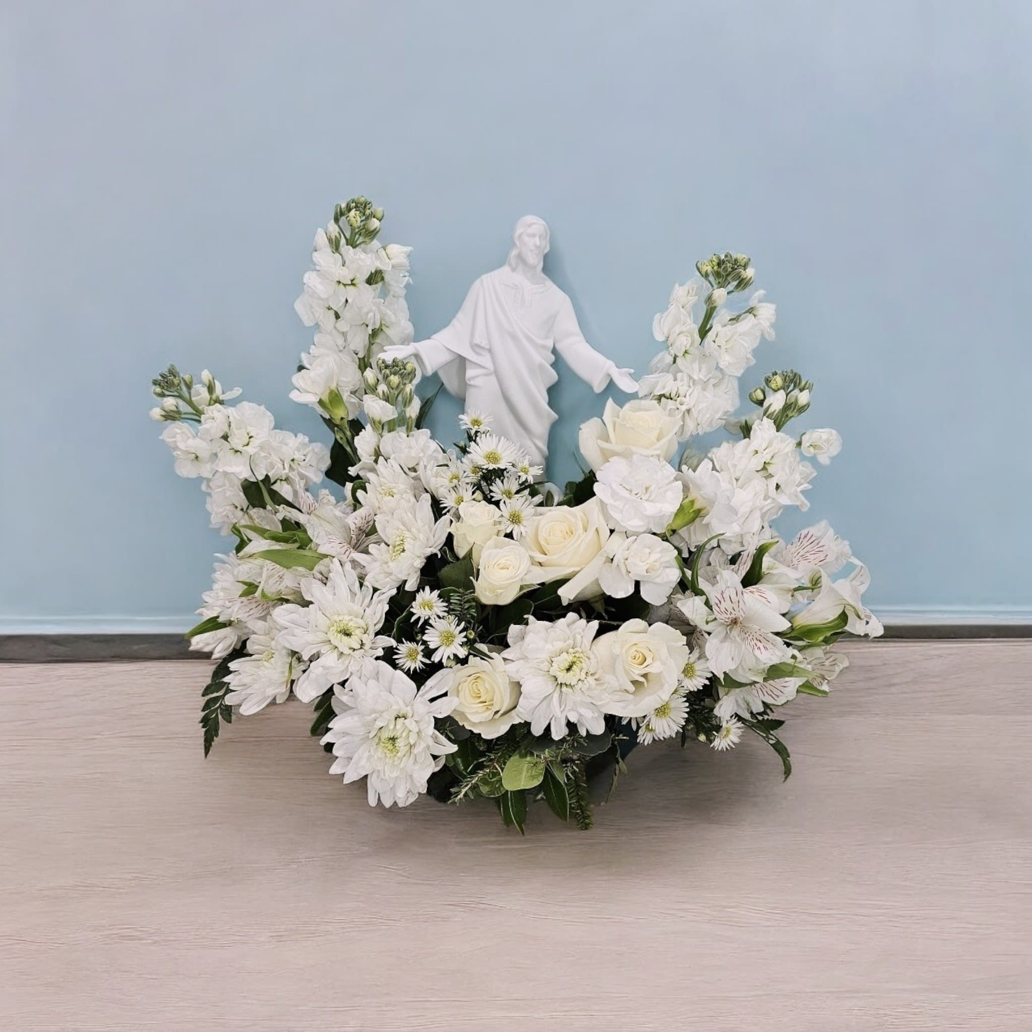 Heavenly Serenity Bouquet - This arrangement reflects the imagery of purity and spirituality with the white roses, stock, cushion, alstroemeria, and mini carnations, all accented by greenery and fine filler. The addition of the sculpture of Jesus in the center adds a meaningful and spiritual touch to the arrangement.