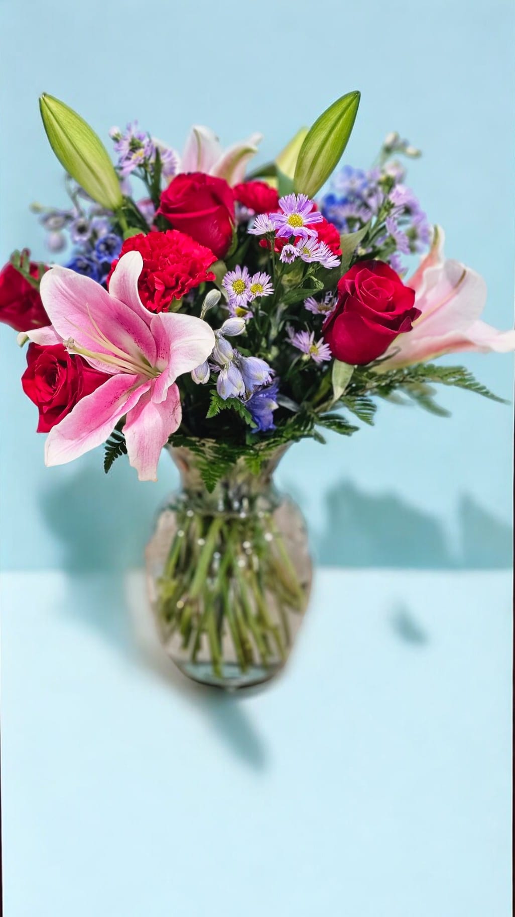 Vivid Elegance - Elevate any space with Vivid Elegance, a stunning arrangement featuring vibrant red roses, delicate blue delphinium, and rich red carnations. Lush purple aster and elegant greenery complement the clear vase, creating a breathtaking display of color and sophistication. Perfect for any occasion.