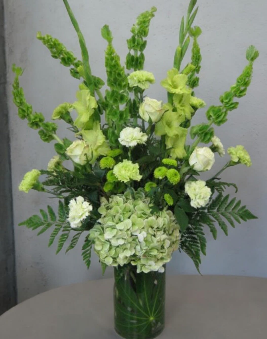 Emerald Elegance - The essence of the arrangement with its predominant green hues (emerald) complemented by the purity of white roses. It also emphasizes the elegance conveyed by the use of an elegant crystal vase, making it sound both sophisticated and visually descriptive. Green Bell Ireland, green Carnations, Green Gladiolus, Green Hydrengas, Green Botton, White Roses, fine Greenery made on fine Vase&nbsp;