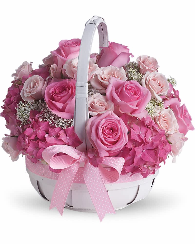 Round basket arrangement of pink roses and hydrangeas with a white handle and pink ribbon bow.