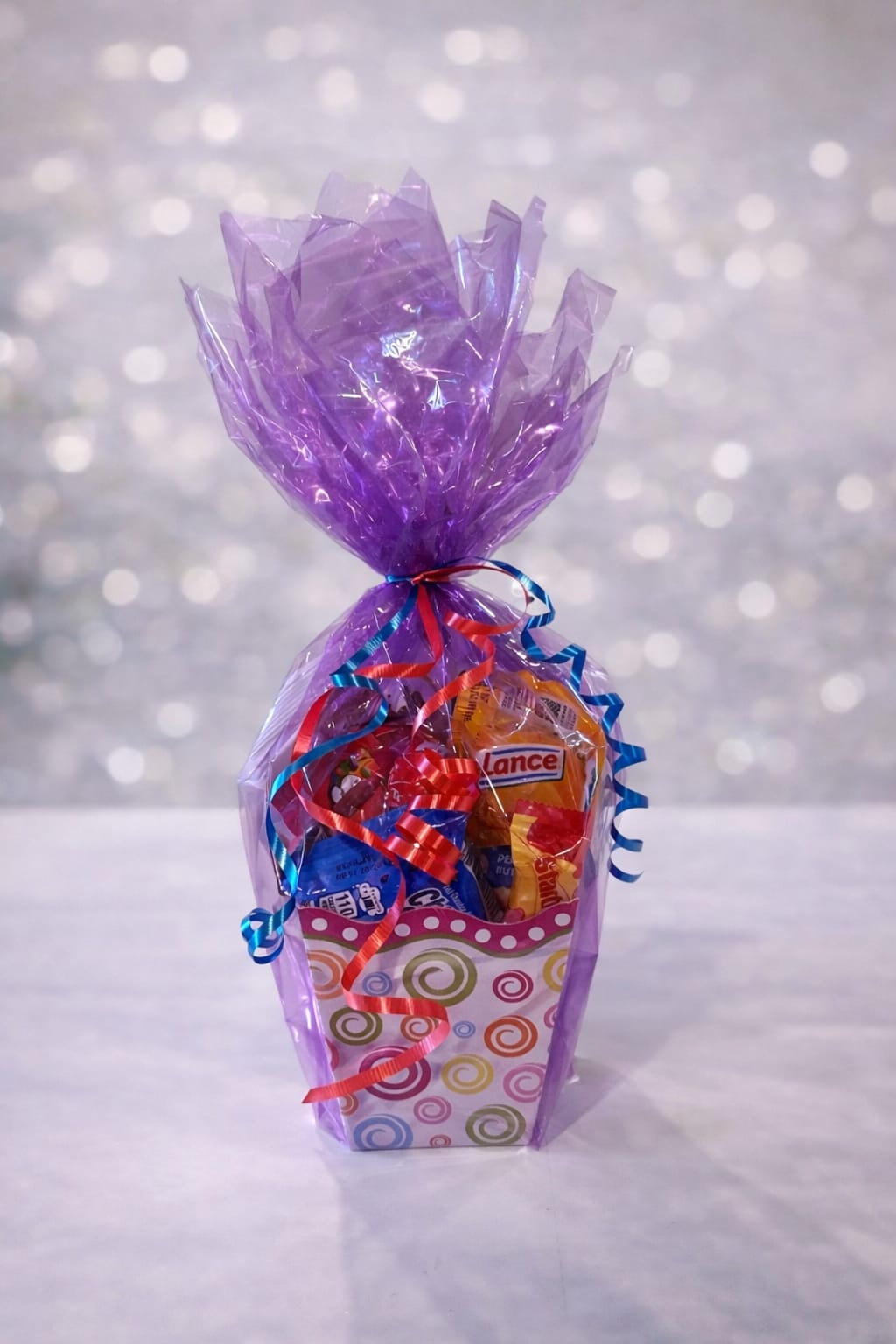 Sweet surprise snack cup - Fun-sized and full of flavor, the Sweet Surprise Snack Cup is a colorful little treat that packs a big smile! Filled with a mix of sweet and savory favorites, this cheerful container is wrapped in vibrant purple cello and finished with playful ribbons for the perfect pop of color. Whether you’re sending a quick pick-me-up, celebrating a birthday, or adding a little extra to a floral delivery, this snack cup is a simple, thoughtful way to brighten someone’s day—because the best surprises come in small packages 
