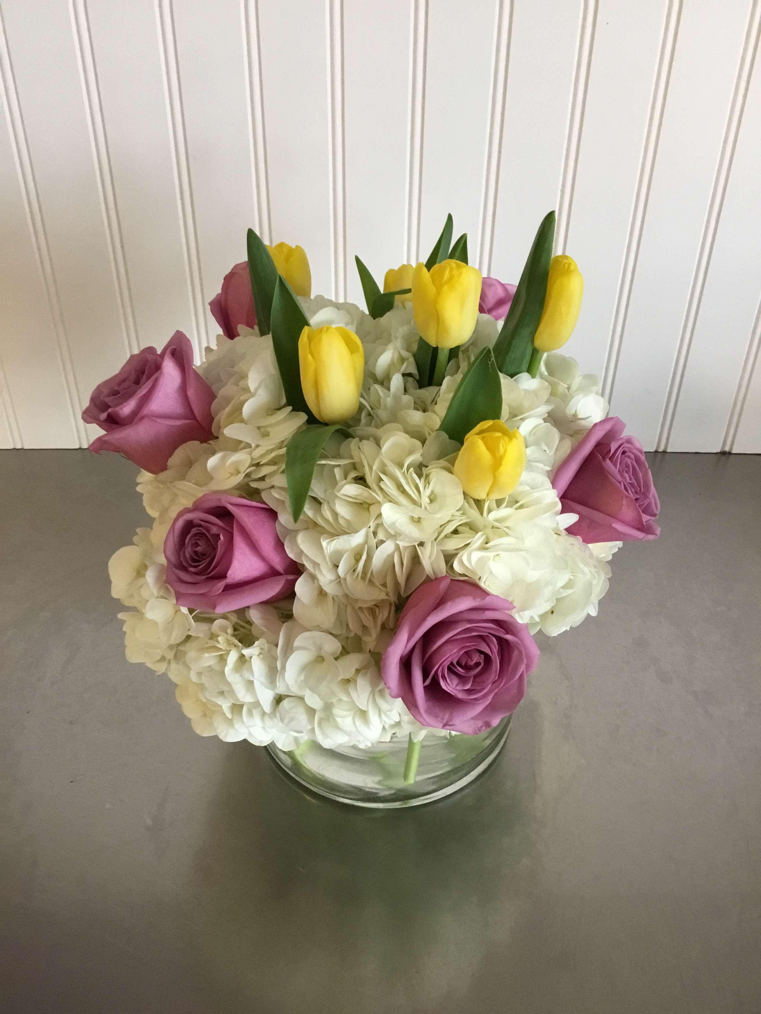 Compact arrangement of yellow tulips, lavender roses, and white hydrangeas in a clear glass vase