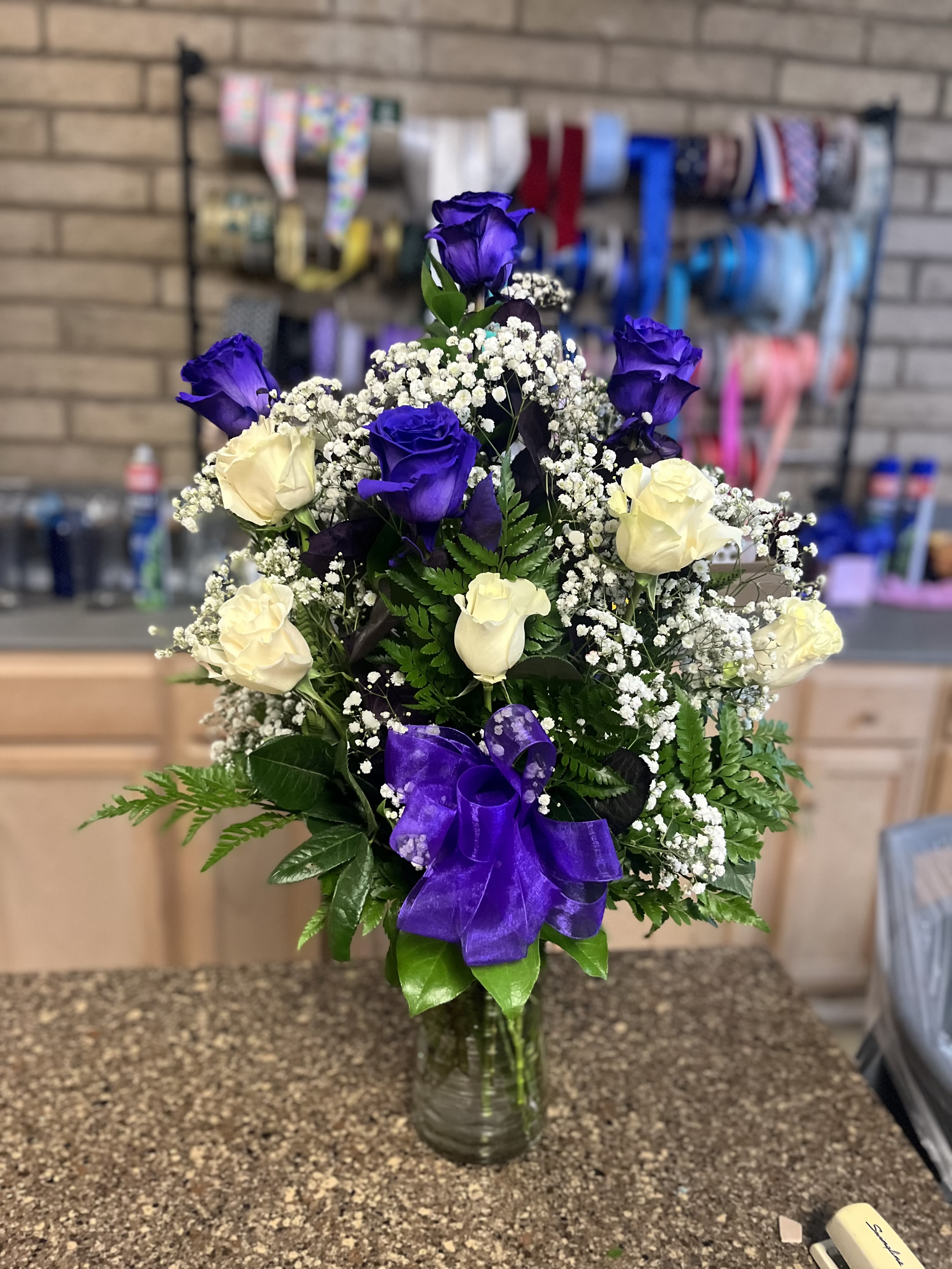 Tall arrangement of purple and white roses with baby's breath in a clear glass vase with a purple bow.