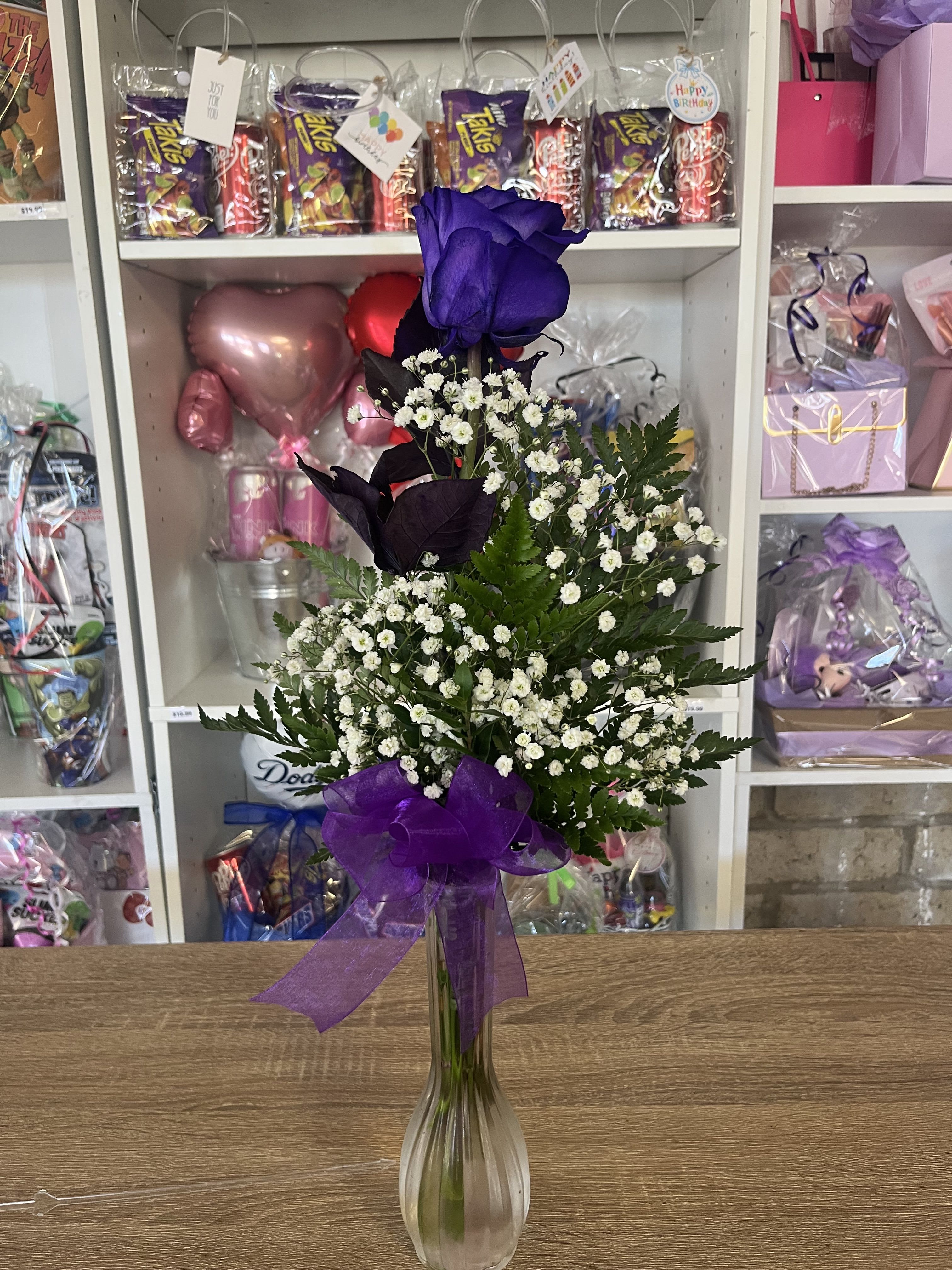 Tall purple rose and white filler flowers in a clear glass vase with a purple bow