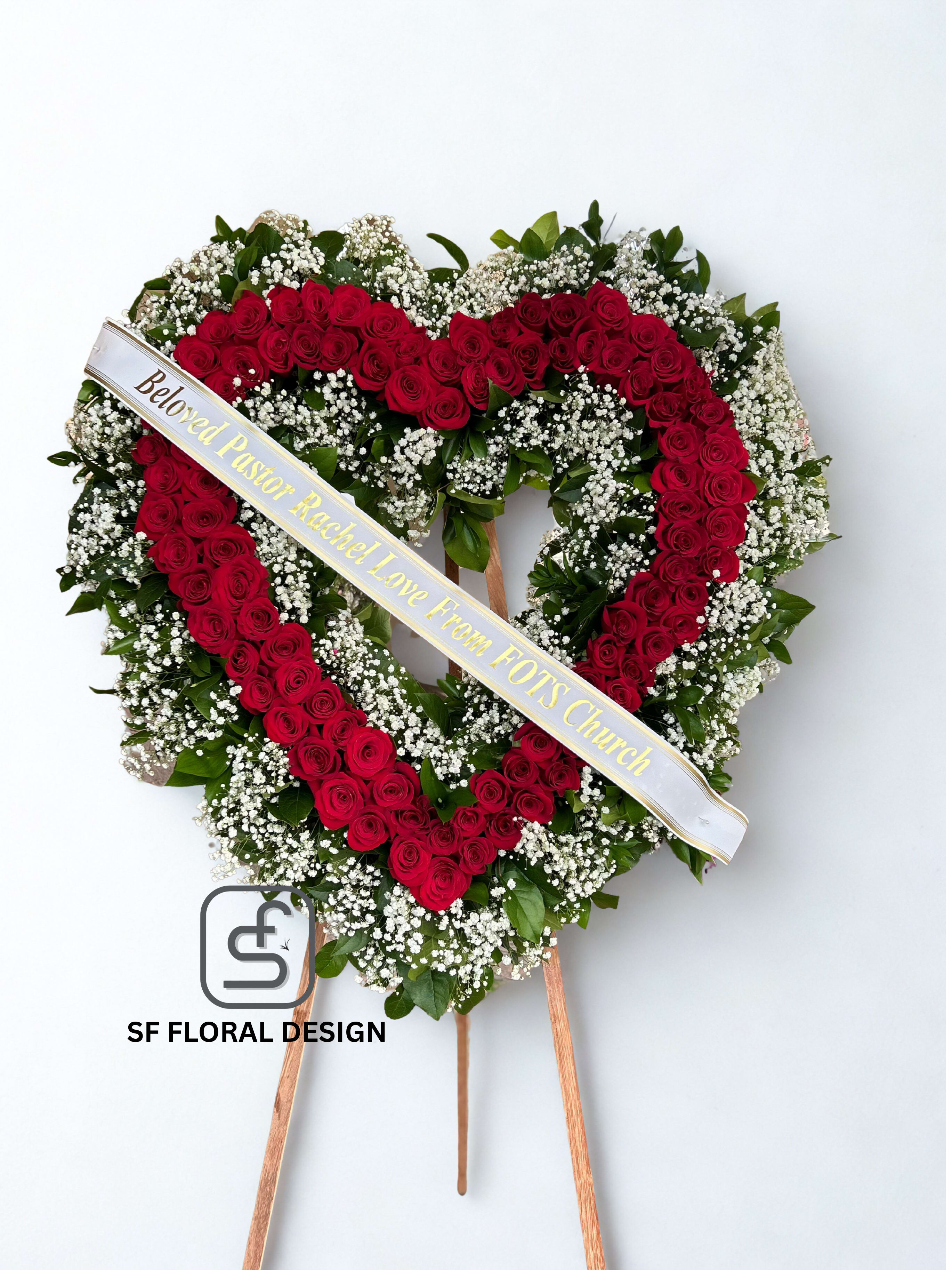 Heart-shaped standing wreath of red roses and white filler flowers with a printed ribbon across the front.