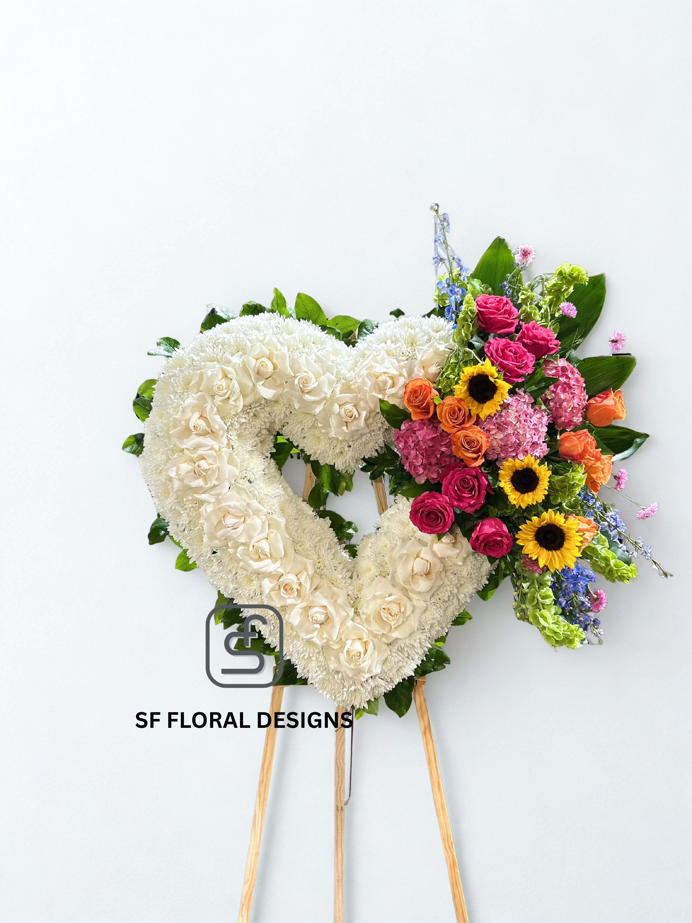 Heart-shaped white standing wreath with colorful roses, sunflowers, and mixed blooms on one side.