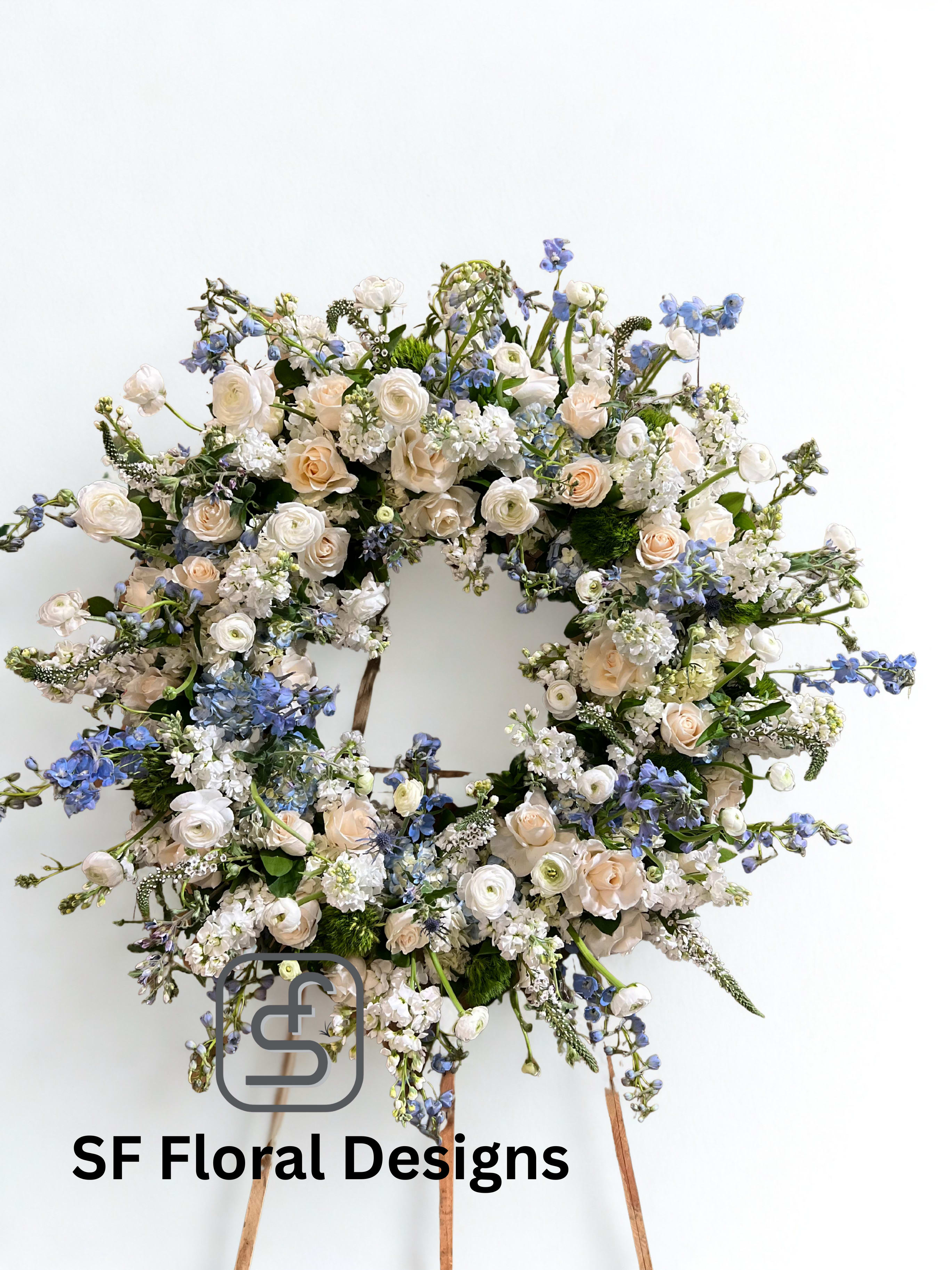 Large round wreath of cream roses and white flowers with soft blue accents on a wooden easel.
