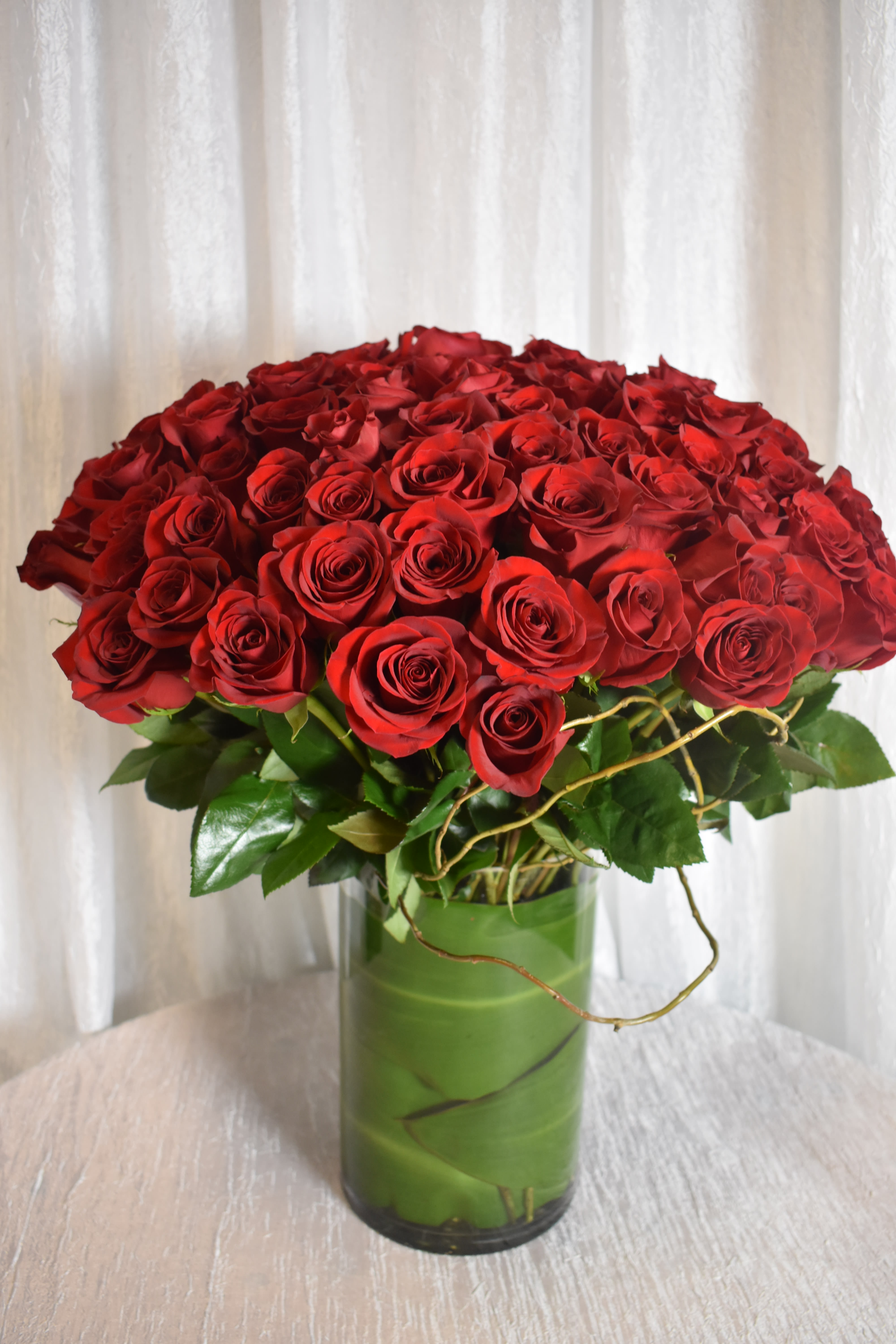 Large arrangement of red roses in a clear cylinder vase wrapped with green leaves.