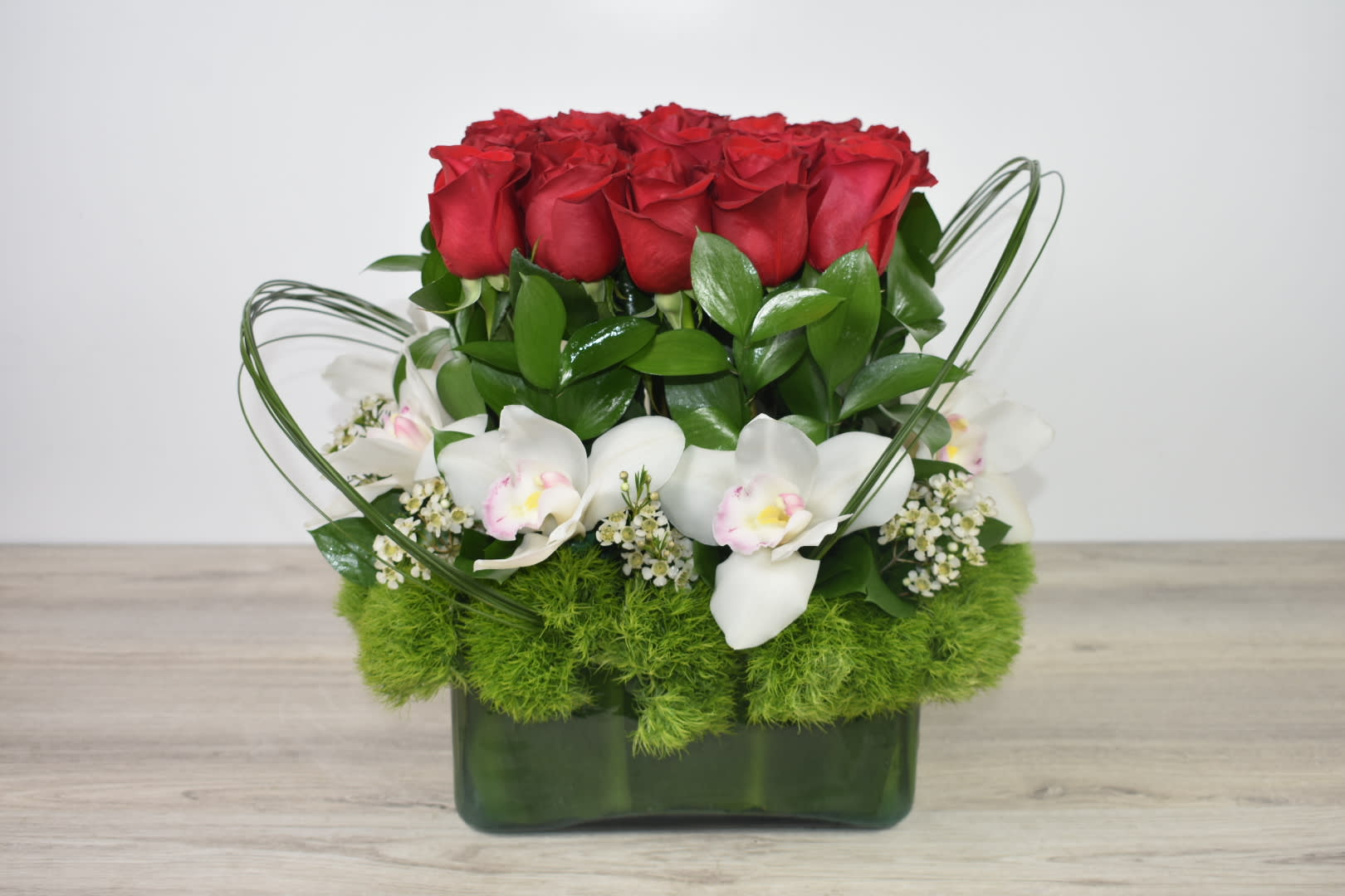 Compact arrangement of red roses, white orchids, and green dianthus in a rectangular green glass vase.