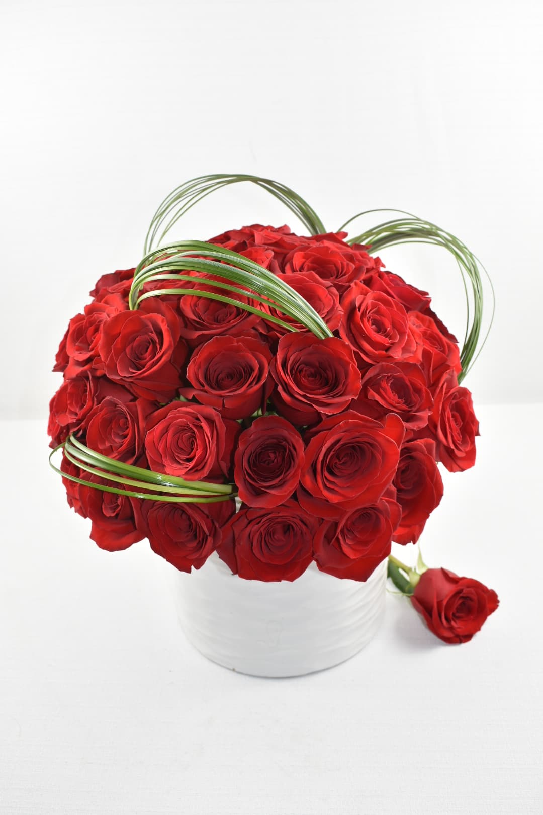 Compact arrangement of red roses in a white vase with looping green blades of grass.