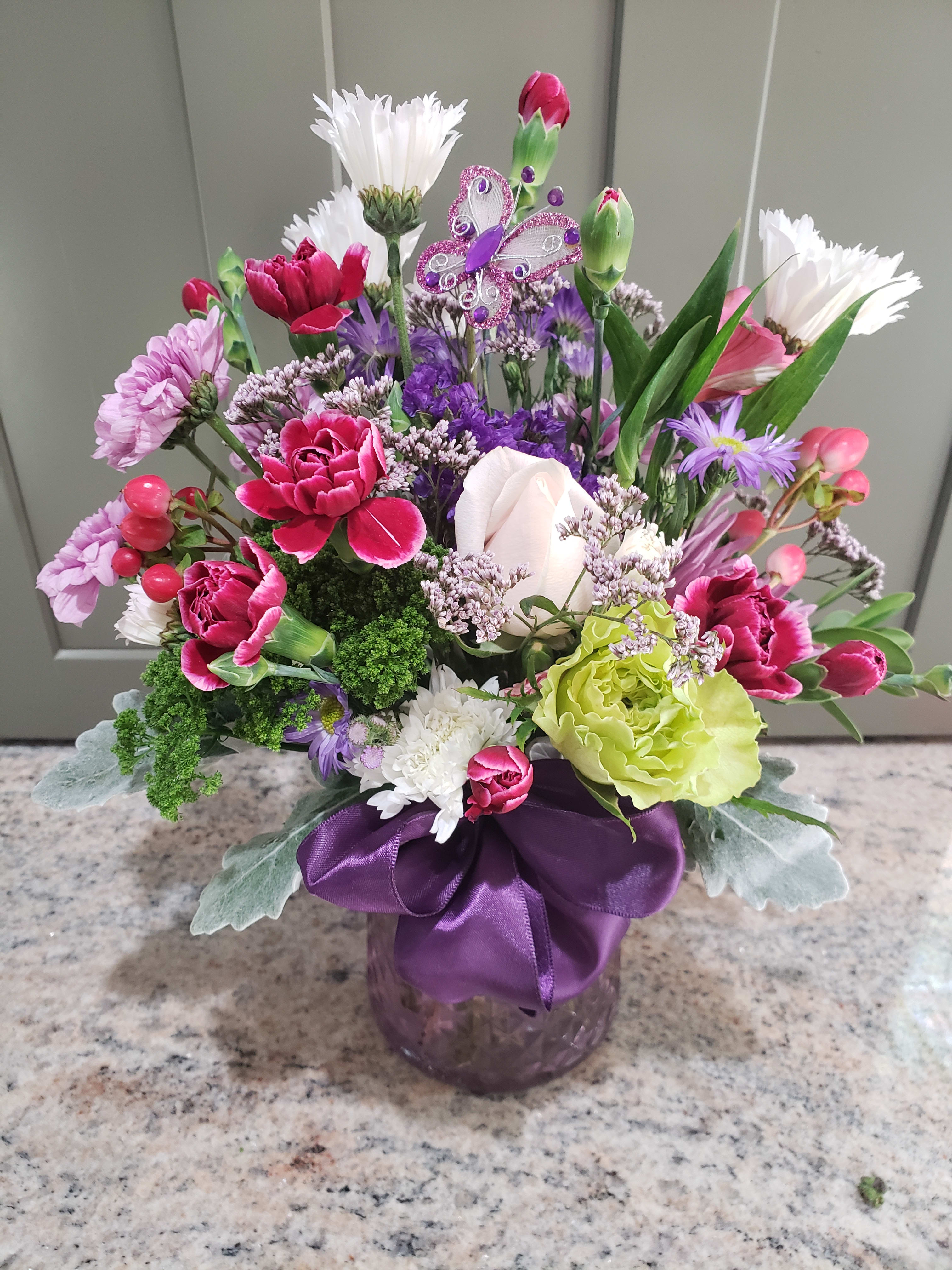 Colorful mixed bouquet of pink, purple, and white flowers in a purple vase with a ribbon and butterfly pick.