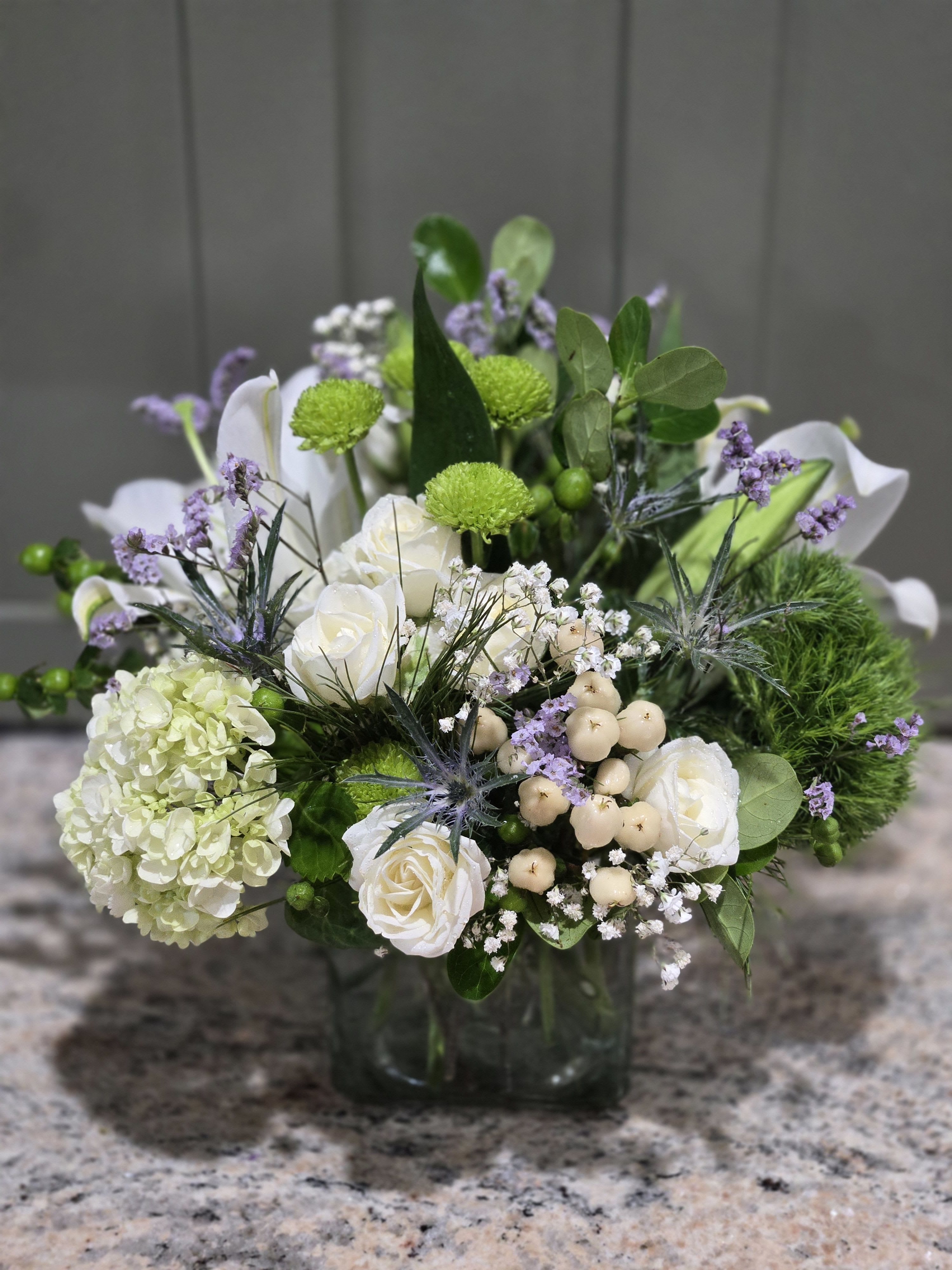 Low arrangement of white roses, hydrangea, green mums, and lavender accents in a glass cube vase