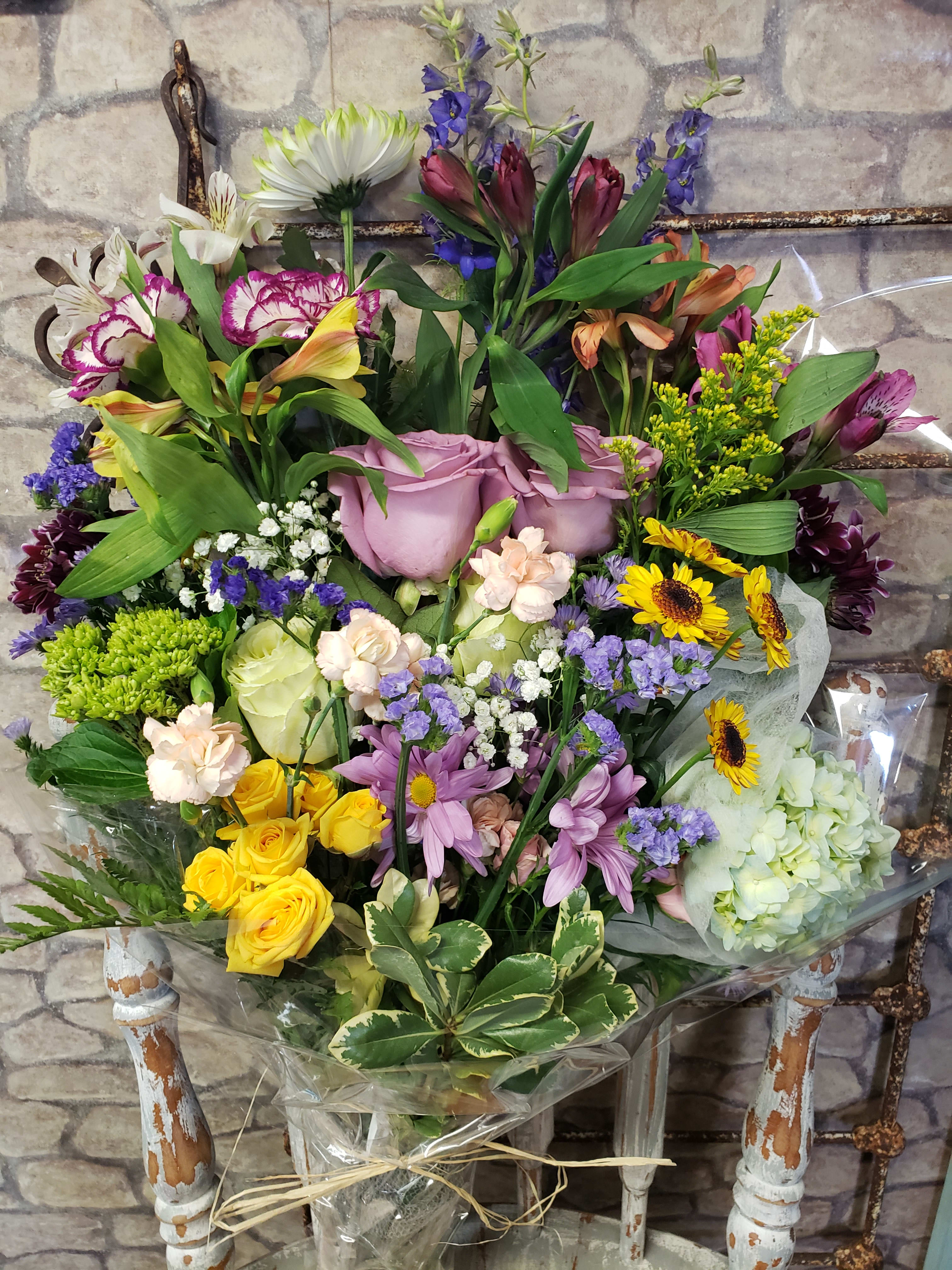 Tall mixed bouquet of roses, daisies, sunflowers and hydrangea wrapped in clear cellophane on a rustic chair