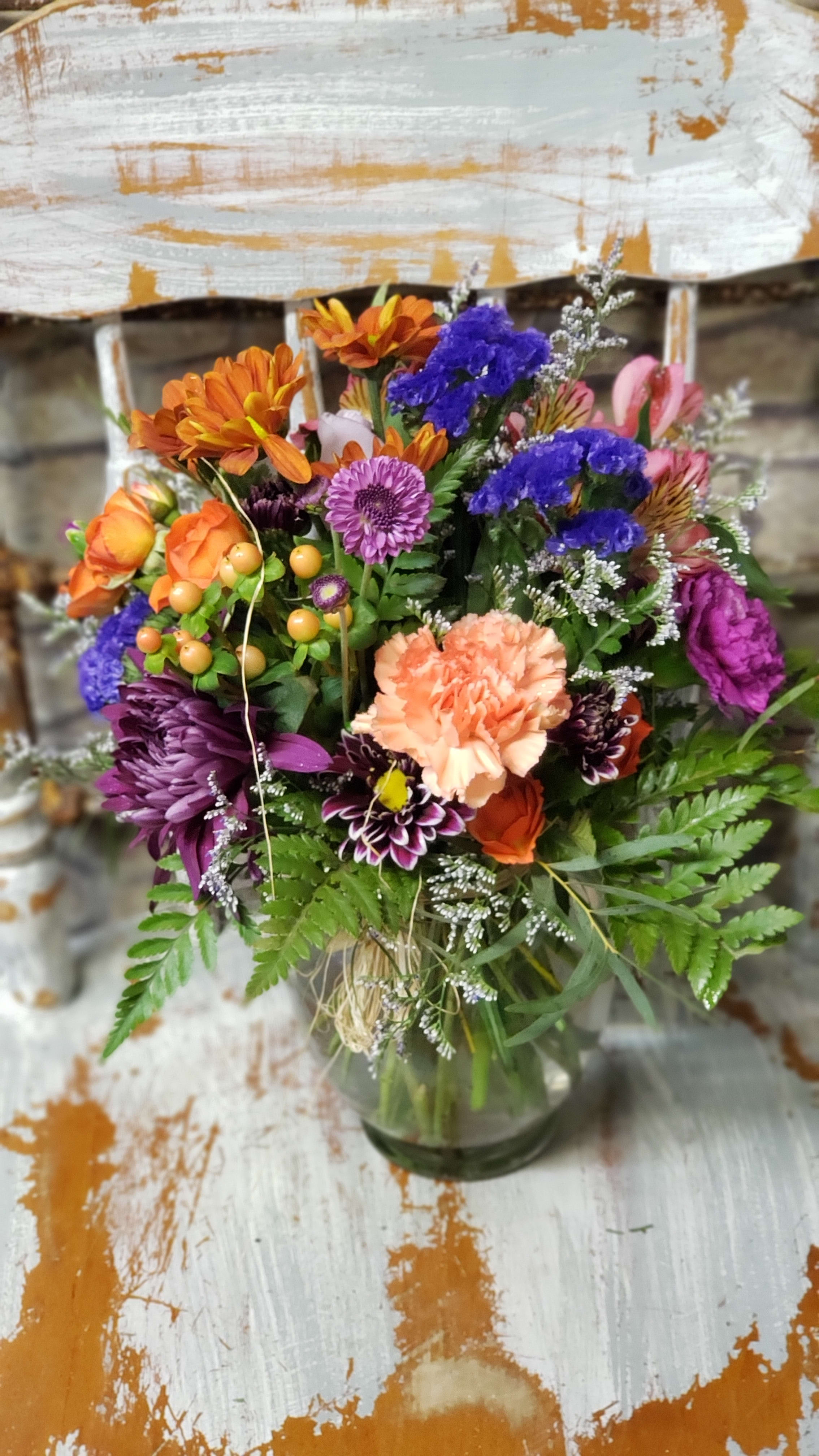 Colorful mixed bouquet with orange roses, peach carnation, and purple blooms in a clear glass vase.
